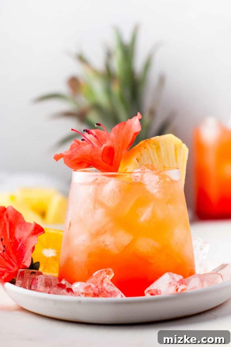 Sunshine Island Rum Punch 2 A beautifully garnished glass of Pineapple Orange Rum Punch with a tropical flower and a fresh pineapple wedge, set against a summer backdrop.