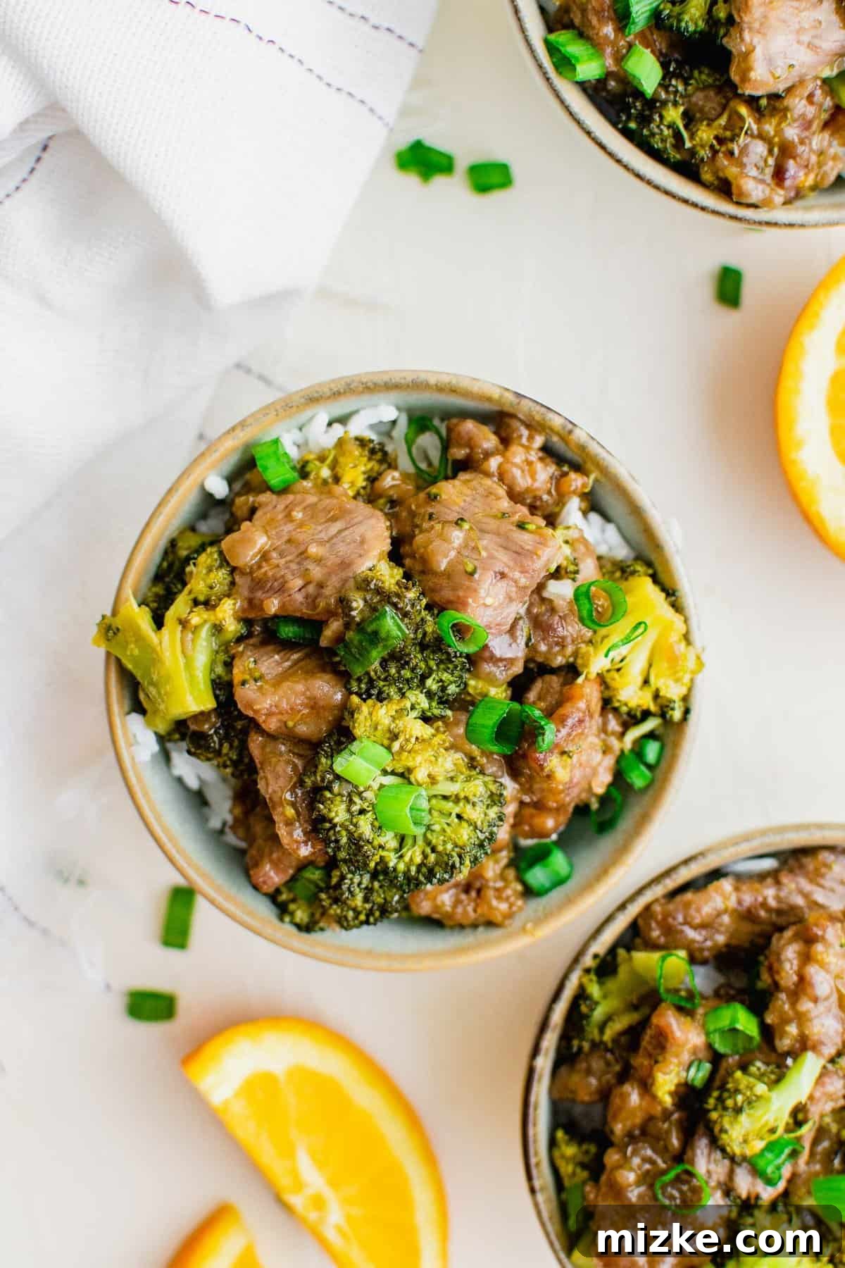 Serve this delightful spicy orange beef with broccoli over rice for a satisfying takeout-style meal that everyone will love. Overhead view of two bowls of orange beef and broccoli served over white rice, garnished with green onions.