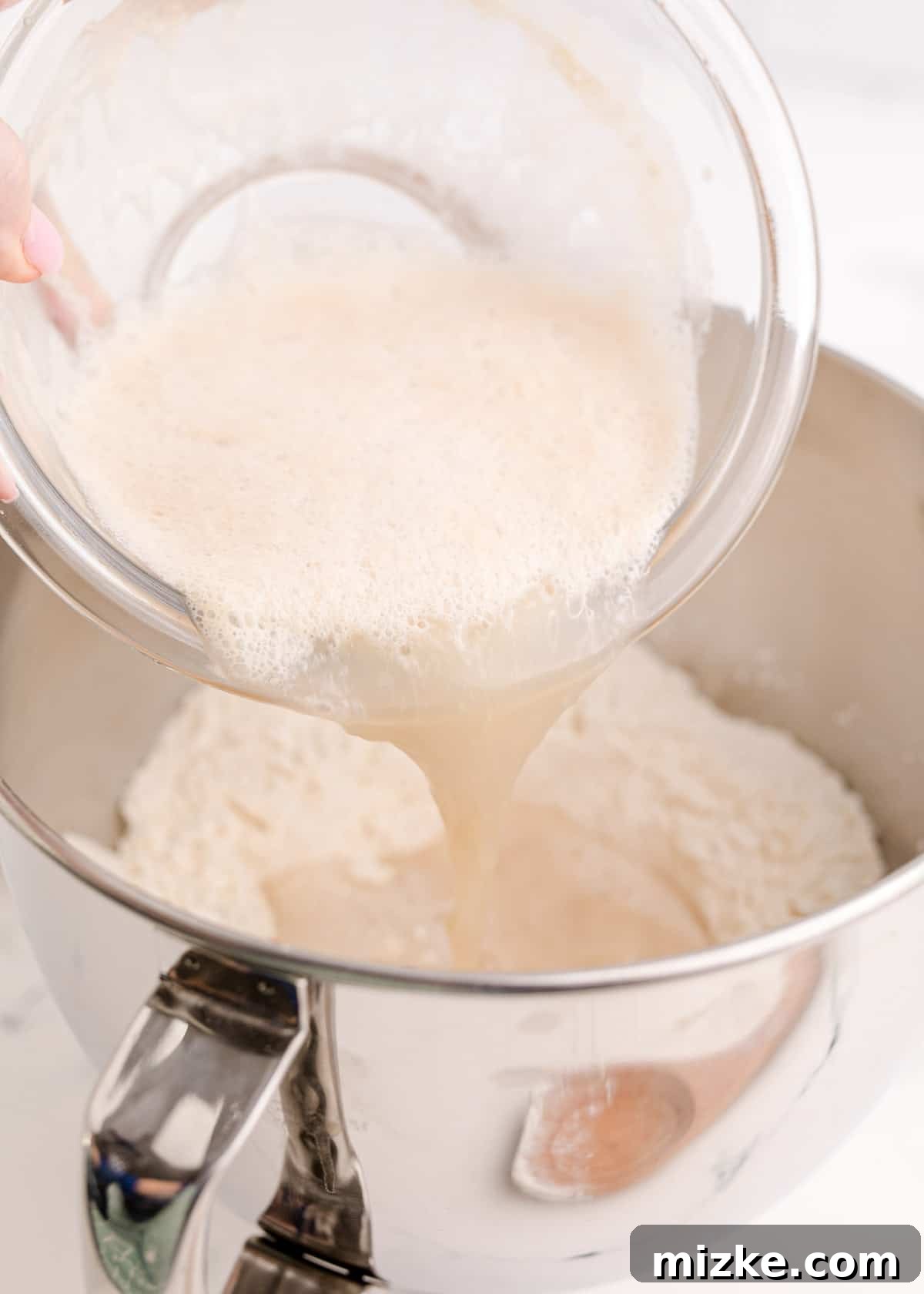 Chewy Homemade Bagels From Scratch 7 The activated yeast mixture being poured into a metal stand mixer bowl containing flour and salt.