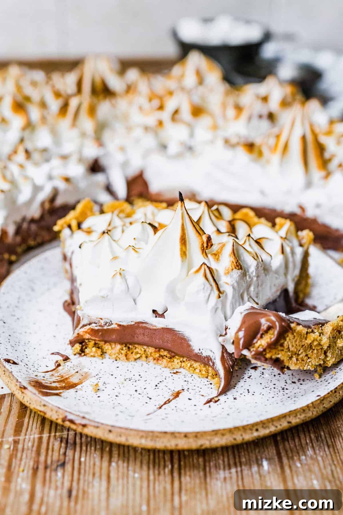 This s'mores tart recipe takes just 15 minutes to make! With a graham cracker crust, ganache filling, and toasty meringue, it's irresistible. A half-eaten slice of s'mores tart on a white plate