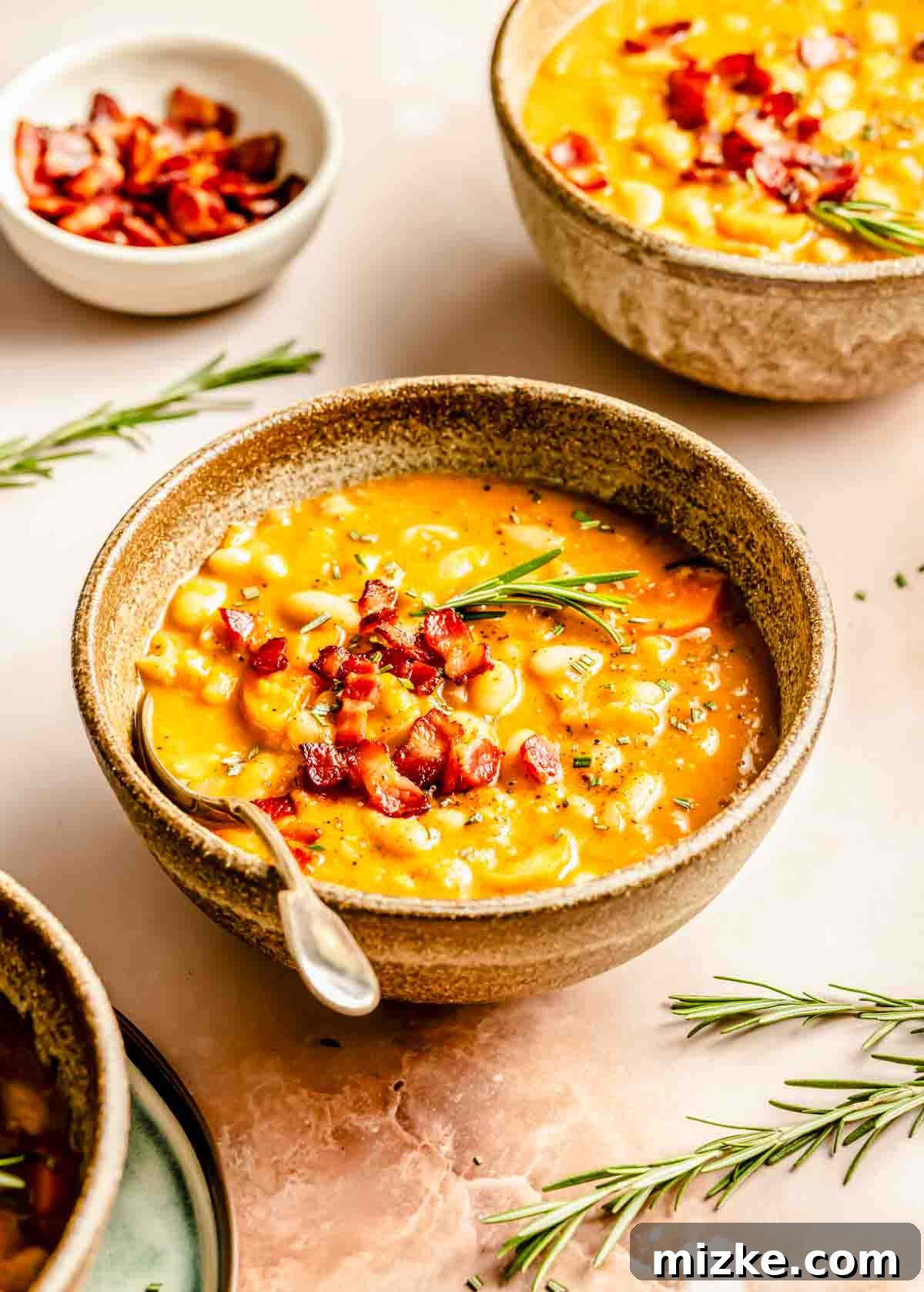 A close-up shot of a bowl of creamy white bean and bacon soup, garnished with crispy bacon bits and a sprig of fresh rosemary. A small bowl of extra sliced bacon sits on the side.