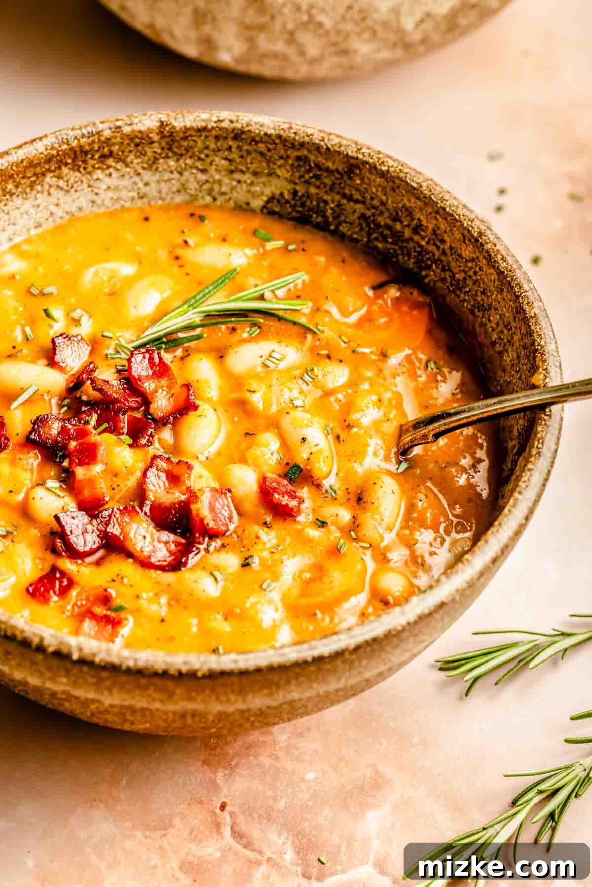 A bowl of rich white bean soup topped with perfectly diced bacon and a sprig of rosemary, with a silver spoon resting on the right side of the bowl.