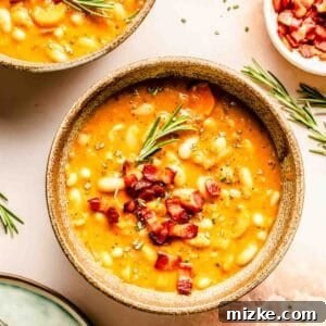 A bowl of white bean soup topped with diced bacon and garnished with rosemary, next to a bowl and spoon.