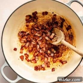 Cooked bacon bits in a large pot with a wooden spoon on a light countertop.