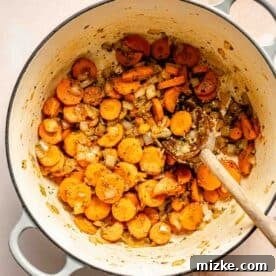 A pot with sliced carrots, onions, and herbs being stirred with a wooden spoon.