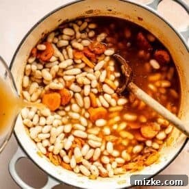 A pot of white bean soup with carrots, being prepared with broth added and a wooden spoon resting inside.