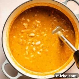 A pot of thick, orange-colored soup with white beans, partially blended with an immersion blender.