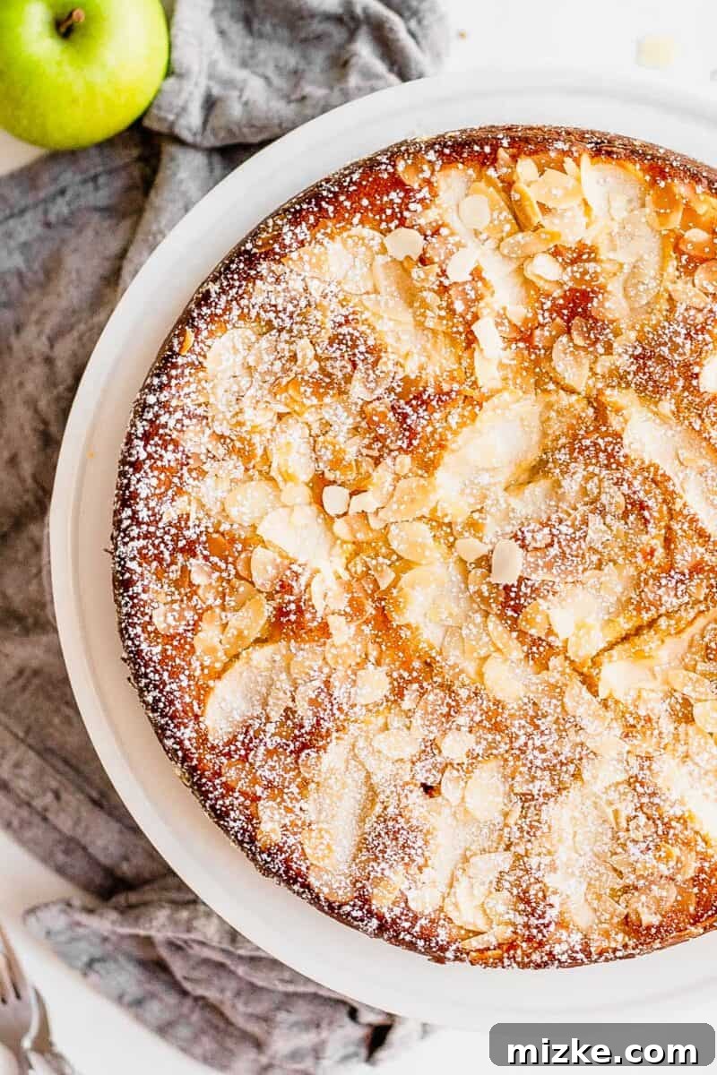 A whole Apple Almond Cake with lightly toasted almonds and sprinkled with powdered sugar sits on a white plate. A gray cloth napkin and whole apple sit next to the cake.