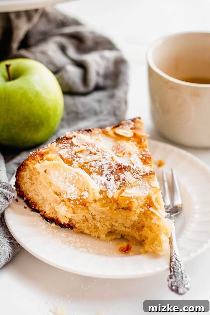 Slice of Apple Almond Cake served on a white dessert plate sits next to a fork. A cup of coffee and a whole apple is in background.