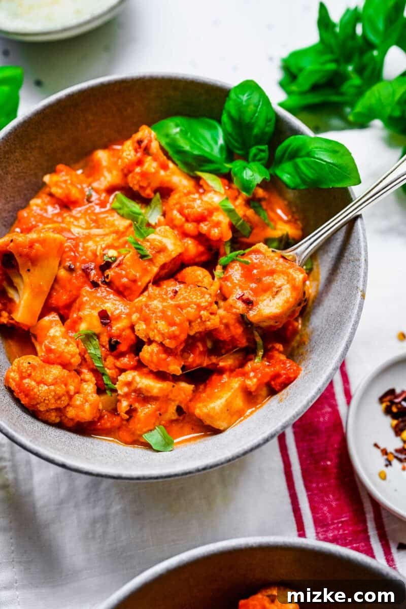 Bite-sized pieces of chicken and cauliflower, coated in marinara and cheese, are being lifted with a fork from the dish.
