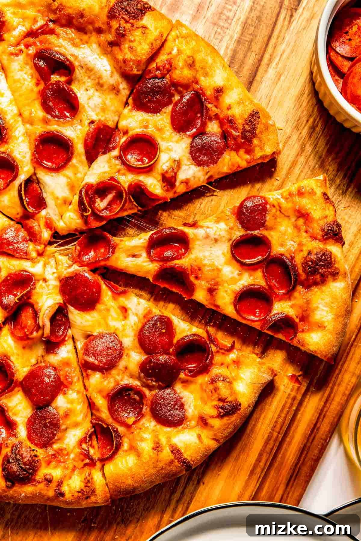 Zesty Pepperoni Pie 3 A close-up shot of a baked and sliced pepperoni pizza resting on a wooden cutting board, with a bowl of fresh pepperoni slices ready on the side for snacking or another pizza.