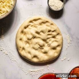 Round pizza dough on a floured surface, surrounded by bowls of shredded cheese, flour, cornmeal, pizza sauce, and sliced pepperoni.