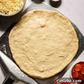 Unbaked pizza dough on a pizza peel surrounded by bowls of shredded cheese, cornmeal, flour, pepperoni slices, and pizza sauce on a white surface.