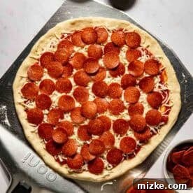 Unbaked pepperoni pizza on a metal pizza peel, surrounded by bowls of cornmeal, flour, pizza sauce, and sliced pepperoni on a white surface.