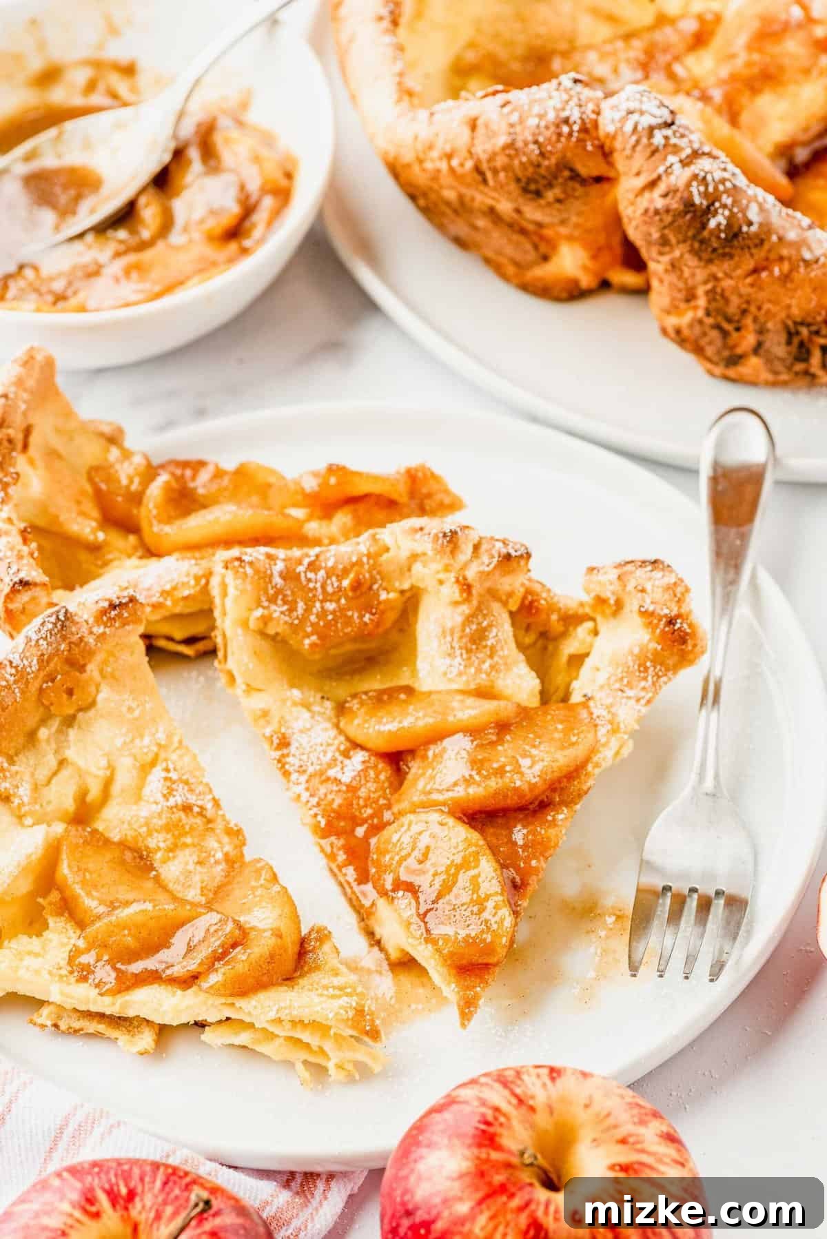 Puffy Apple Cinnamon Dutch Baby 9 Several slices of golden Apple Cinnamon Dutch Baby Pancake, topped with rich apple compote, are beautifully arranged and ready to be served on a plate. A fork rests beside them.