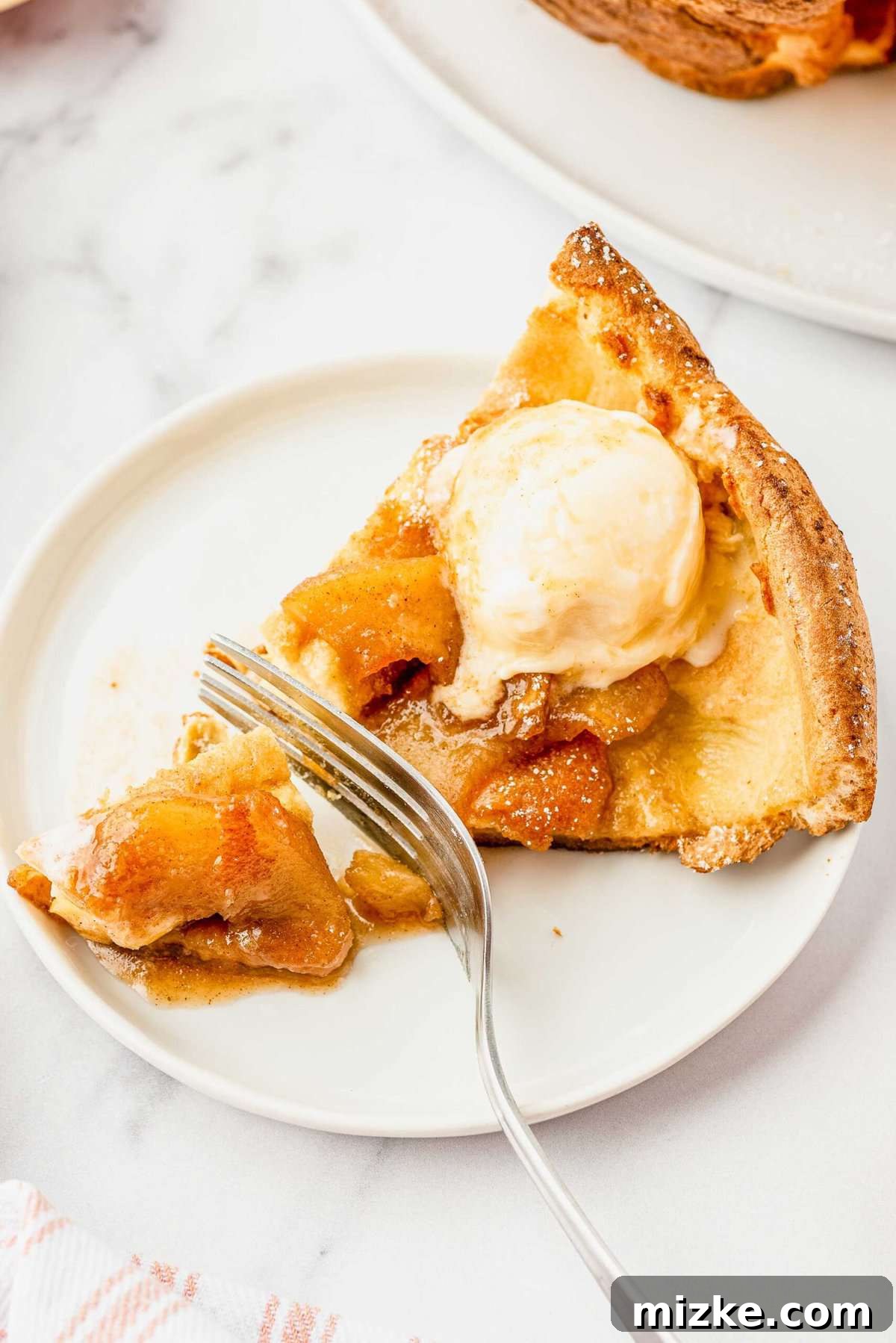 Puffy Apple Cinnamon Dutch Baby 10 A fork cuts through a single fluffy slice of Apple Cinnamon Dutch Baby Pancake on a white plate, revealing its custardy interior and showing the readiness to enjoy.