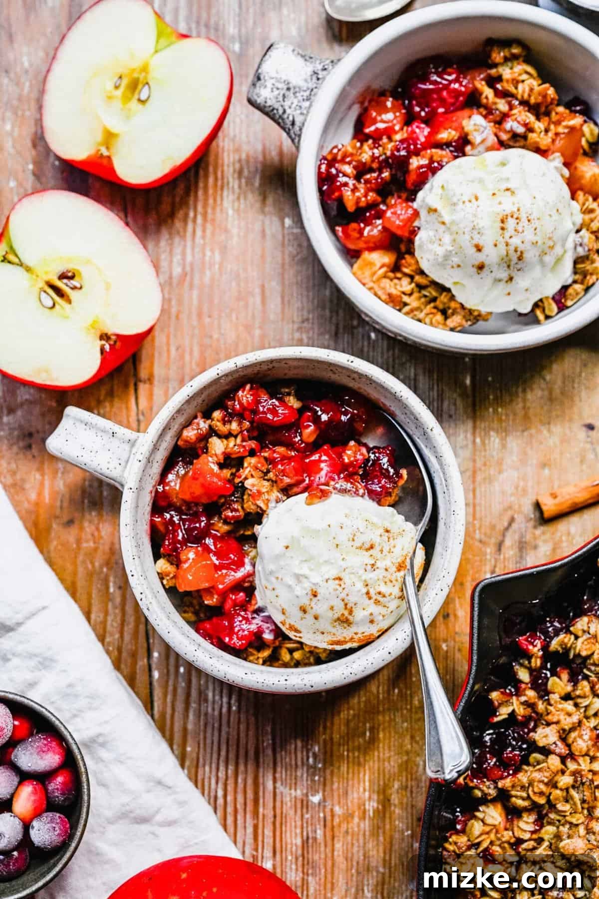 Serve individual portions of Apple Cranberry Crisp with a scoop of ice cream for a delightful dessert. Individual servings of warm apple cranberry crisp with vanilla ice cream, presented in white mugs on a rustic wooden surface, alongside a sliced apple.