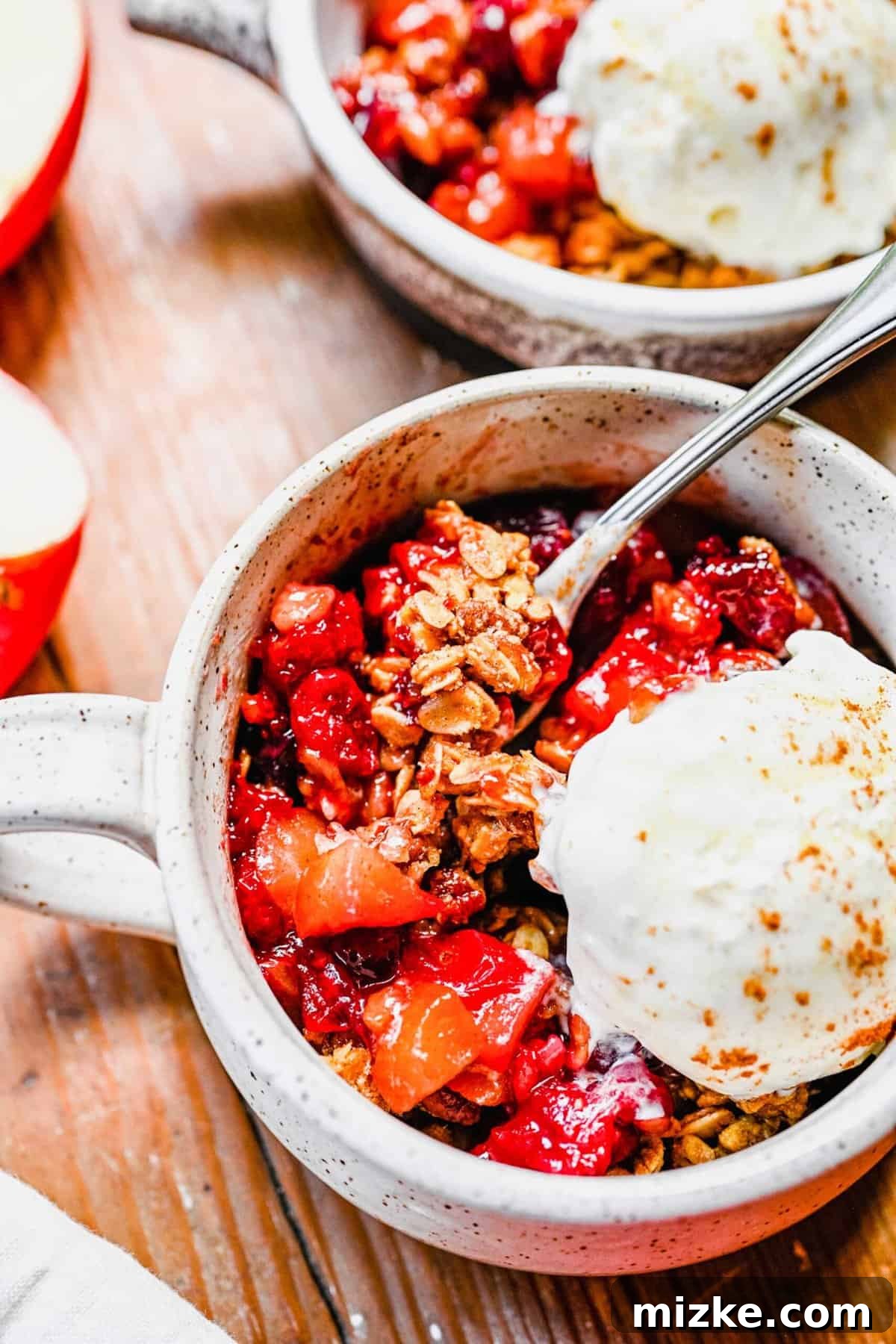 Enjoy warm Apple Cranberry Crisp with a scoop of ice cream. A single serving of golden-brown apple cranberry crisp in a white mug, topped with a generous scoop of vanilla ice cream.