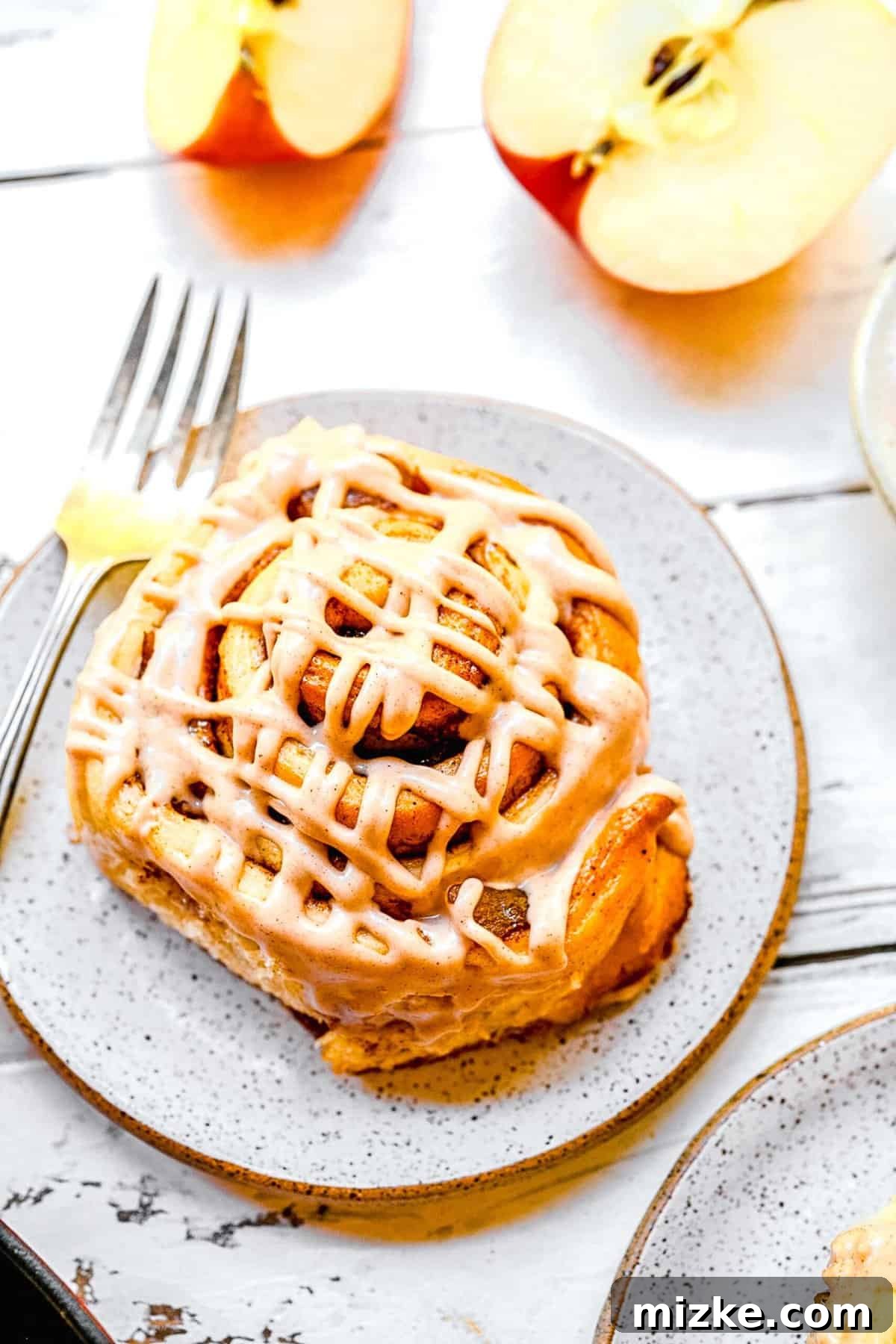 A single, beautifully frosted apple pie cinnamon roll resting on an elegant gold-rimmed dessert plate with a fork.