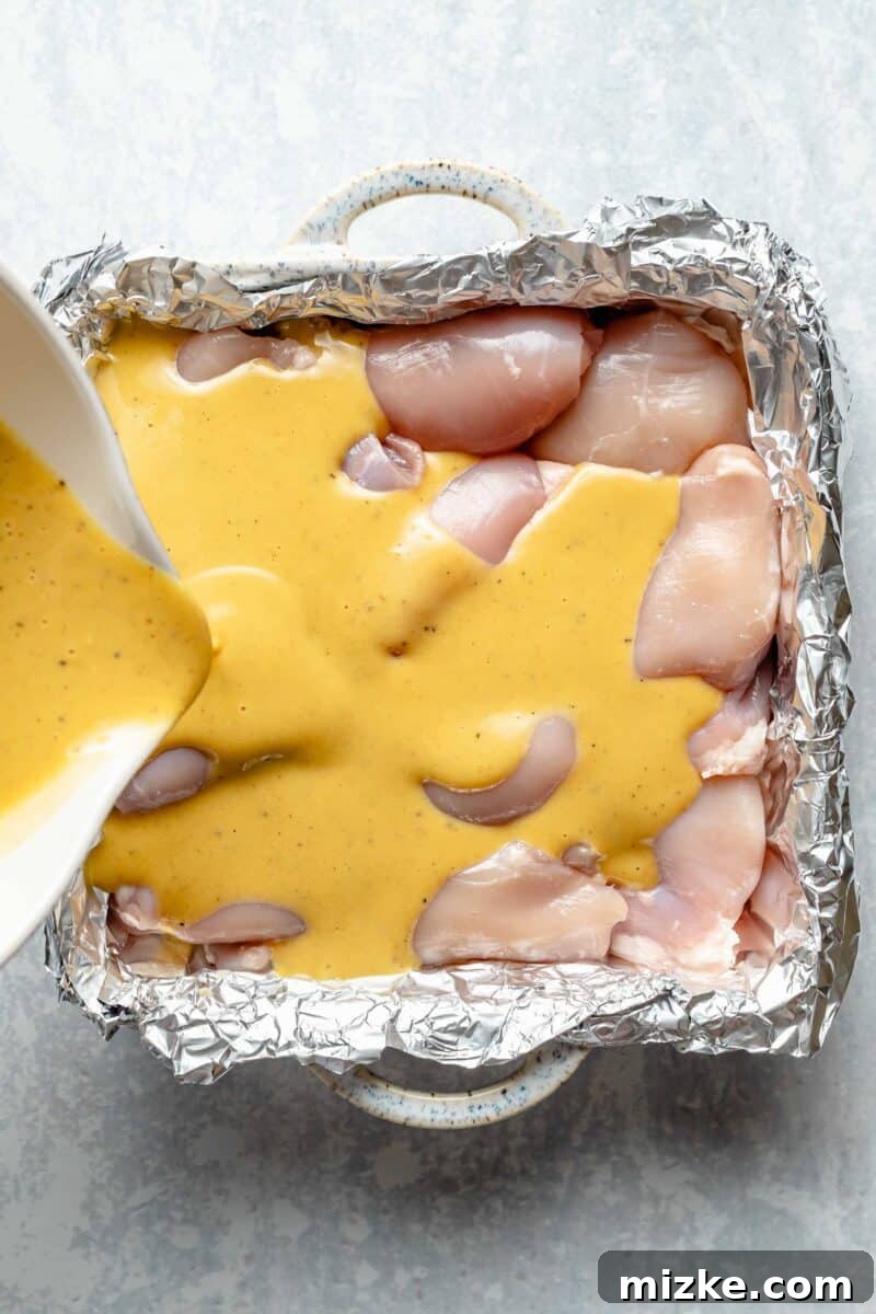 Coating boneless, skinless chicken thighs generously with the delicious maple-Dijon sauce before baking. Raw chicken thighs in a foil-lined baking dish, being covered with the maple-Dijon sauce.