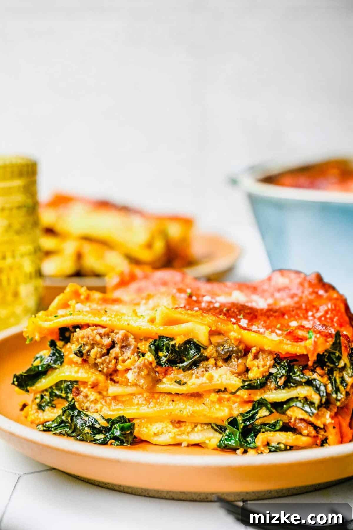 Creamy Pumpkin Lasagna with Savory Sausage and Fresh Kale - A Delightful Autumn Dish Close up of a slice of pumpkin lasagna on a plate, with another slice and the baking dish in the background, showcasing its rich layers and golden cheese