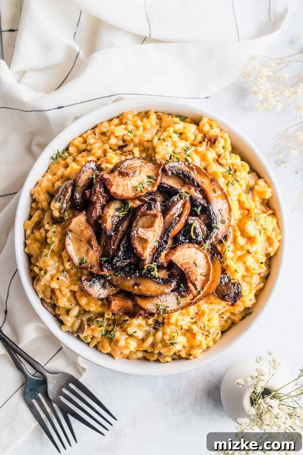 Creamy and healthy vegan mushroom risotto, a perfect plant-based fall dinner. An overhead view of a beautifully presented serving bowl of vegan mushroom risotto, adorned with roasted mushrooms and fresh herbs, arranged alongside elegant utensils, a textured kitchen towel, and a vase of flowers, creating a rustic yet refined dining scene.