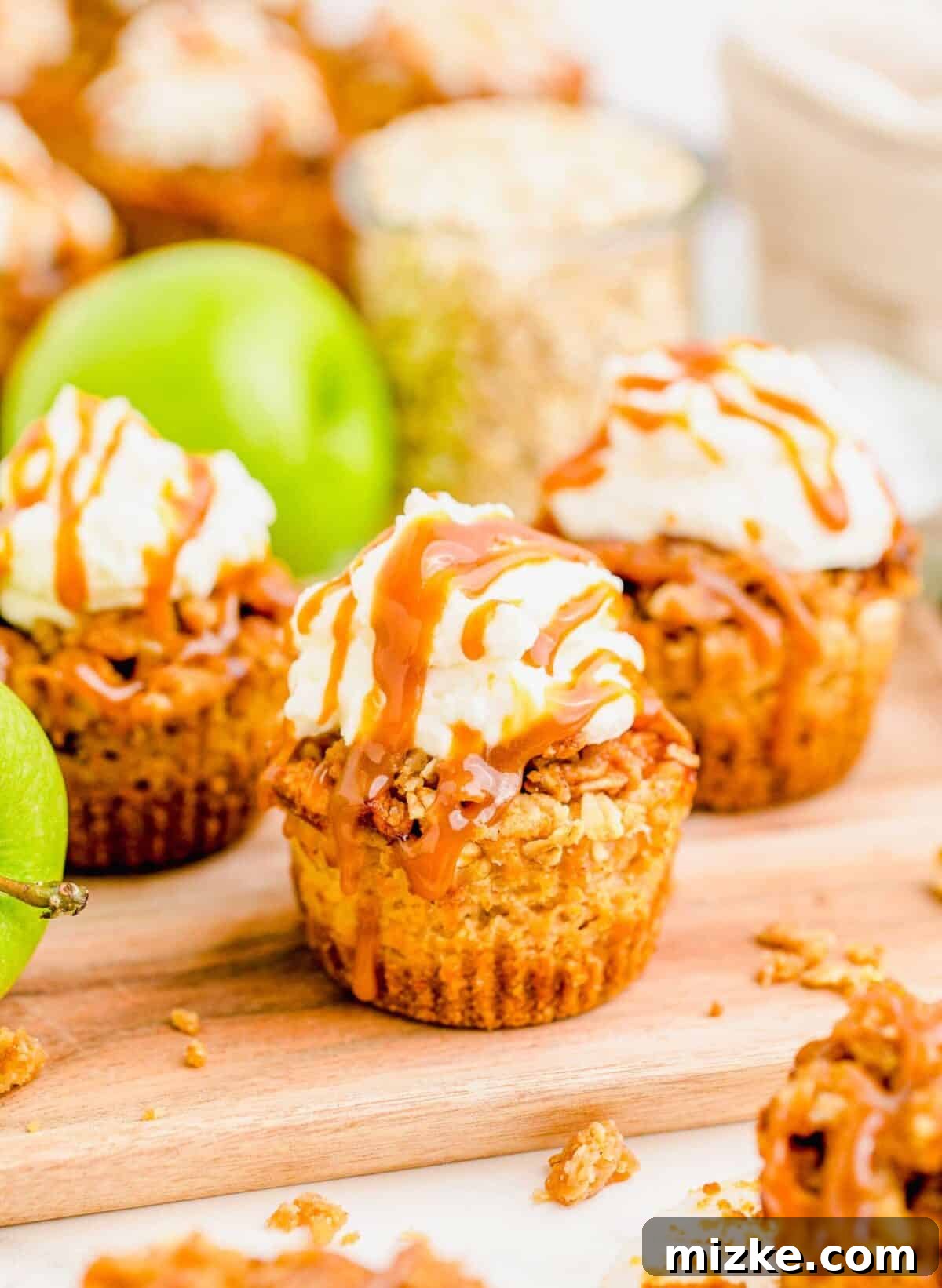 Mini Caramel Apple Cheesecakes: A delightful fall dessert with a crunchy graham cracker crust, creamy filling, spiced apples, and a crumbly oat topping. Close-up of three beautifully arranged mini caramel apple cheesecakes, with a fresh green apple and a rustic jar of oats in the softly blurred background, highlighting the key ingredients and the dessert's aesthetic appeal.