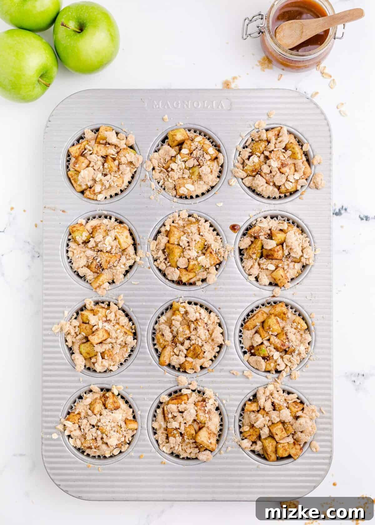 Muffin tray filled with unbaked mini caramel apple cheesecakes. A fully assembled 12-cup muffin tray, showcasing unbaked mini caramel apple cheesecakes. Each cup holds a graham cracker crust, a creamy filling, a layer of spiced apples, and a generous streusel topping, all ready for the oven.