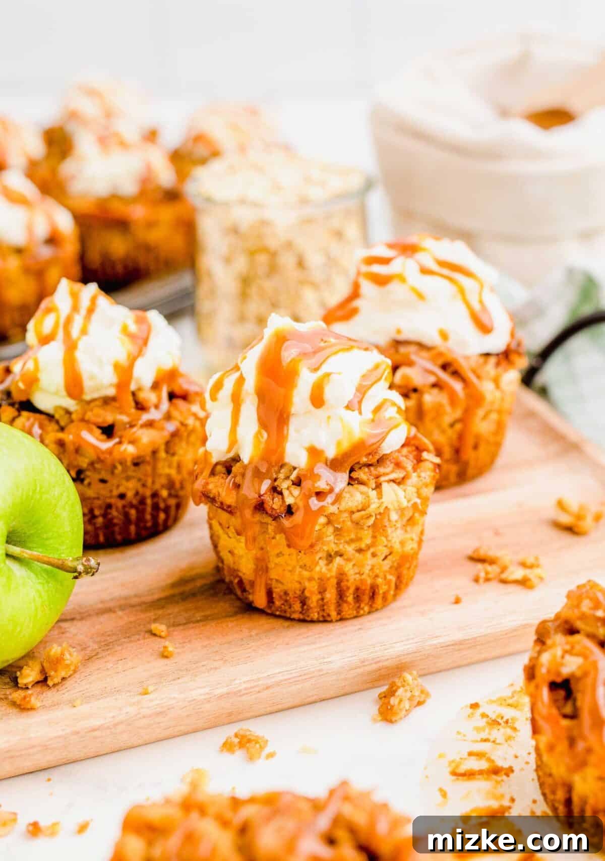 Three mini caramel apple cheesecakes with an apple and oats in the background. A close-up of three exquisite mini caramel apple cheesecakes, artfully arranged next to a crisp red apple. A rustic jar of rolled oats provides a cozy backdrop, emphasizing the wholesome and autumnal essence of the dessert.