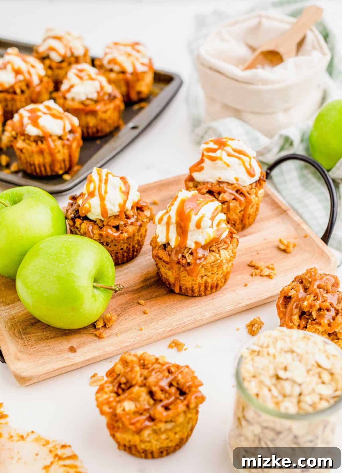 Three mini caramel apple cheesecakes on a cutting board with apples. A beautifully composed shot featuring three mini caramel apple cheesecakes resting on a rustic cutting board, flanked by two fresh Granny Smith apples. More cheesecakes are softly blurred in the background, creating an inviting and abundant scene.
