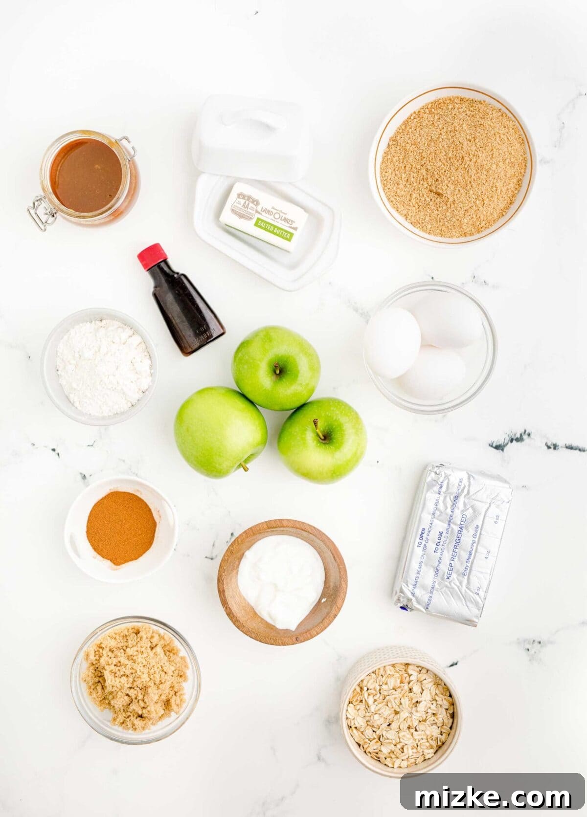 Gather your ingredients for these delightful mini caramel apple cheesecakes: fresh apples, cream cheese, graham crumbs, oats, butter, eggs, and spices for a homemade fall treat. Overhead view of all the fresh ingredients needed for baking mini caramel apple cheesecakes, including vibrant green apples, rich cream cheese, vanilla extract, graham cracker crumbs, rolled oats, butter, eggs, flour, and various spices arranged beautifully on a rustic wooden surface.