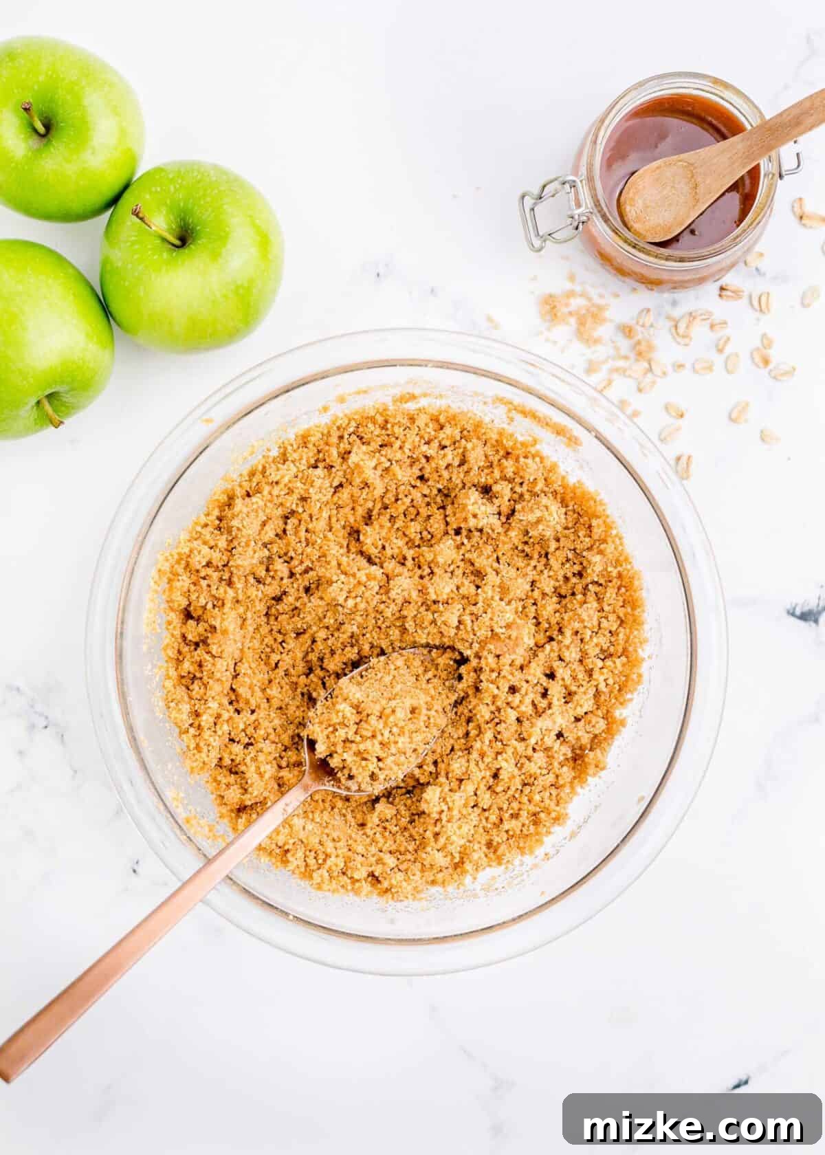 Ingredients for the crumble topping: flour, brown sugar, rolled oats, cinnamon, butter, with apples on the side. A mixing bowl filled with the dry and wet ingredients for the crunchy oat topping: flour, brown sugar, rolled oats, cinnamon, and chunks of butter, alongside three fresh apples, ready to be combined.