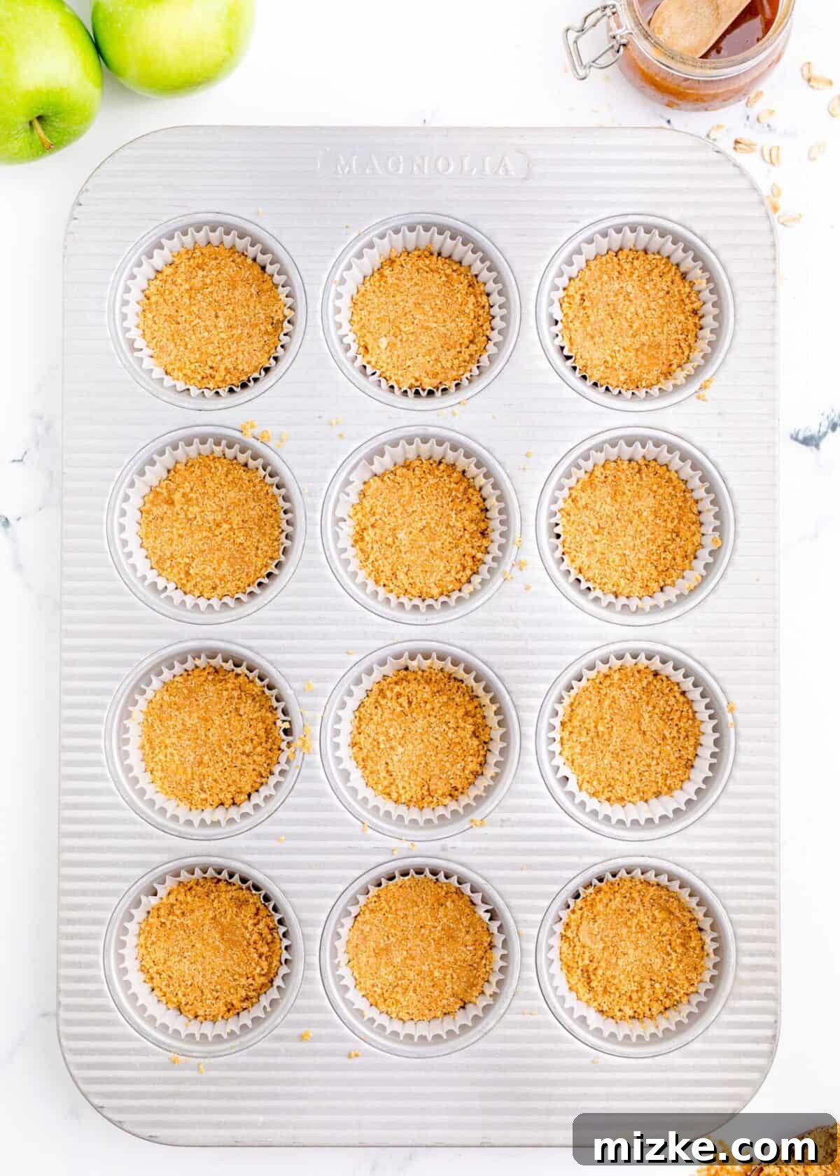 Muffin tray with graham cracker crusts in each liner, prepared for baking. A 12-cup muffin tin meticulously lined with paper liners, each containing a perfectly pressed graham cracker crust base, ready for the creamy cheesecake filling.
