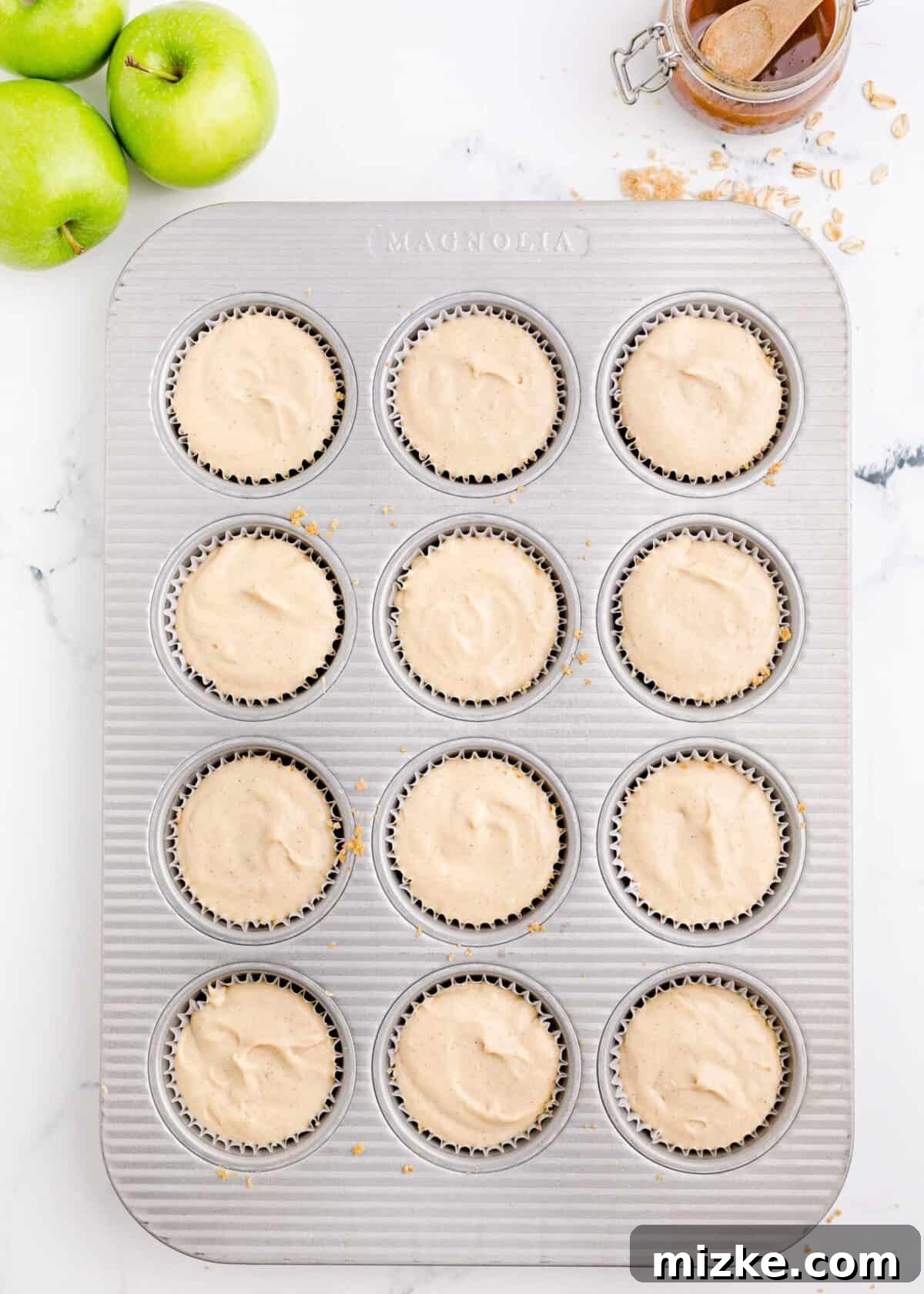 Muffin tray filled with uncooked cheesecake batter over the crusts. A standard 12-cup muffin tray, with each cup filled to an even level with the uncooked, creamy cheesecake batter, set atop its graham cracker crust, ready for the apple and streusel toppings.