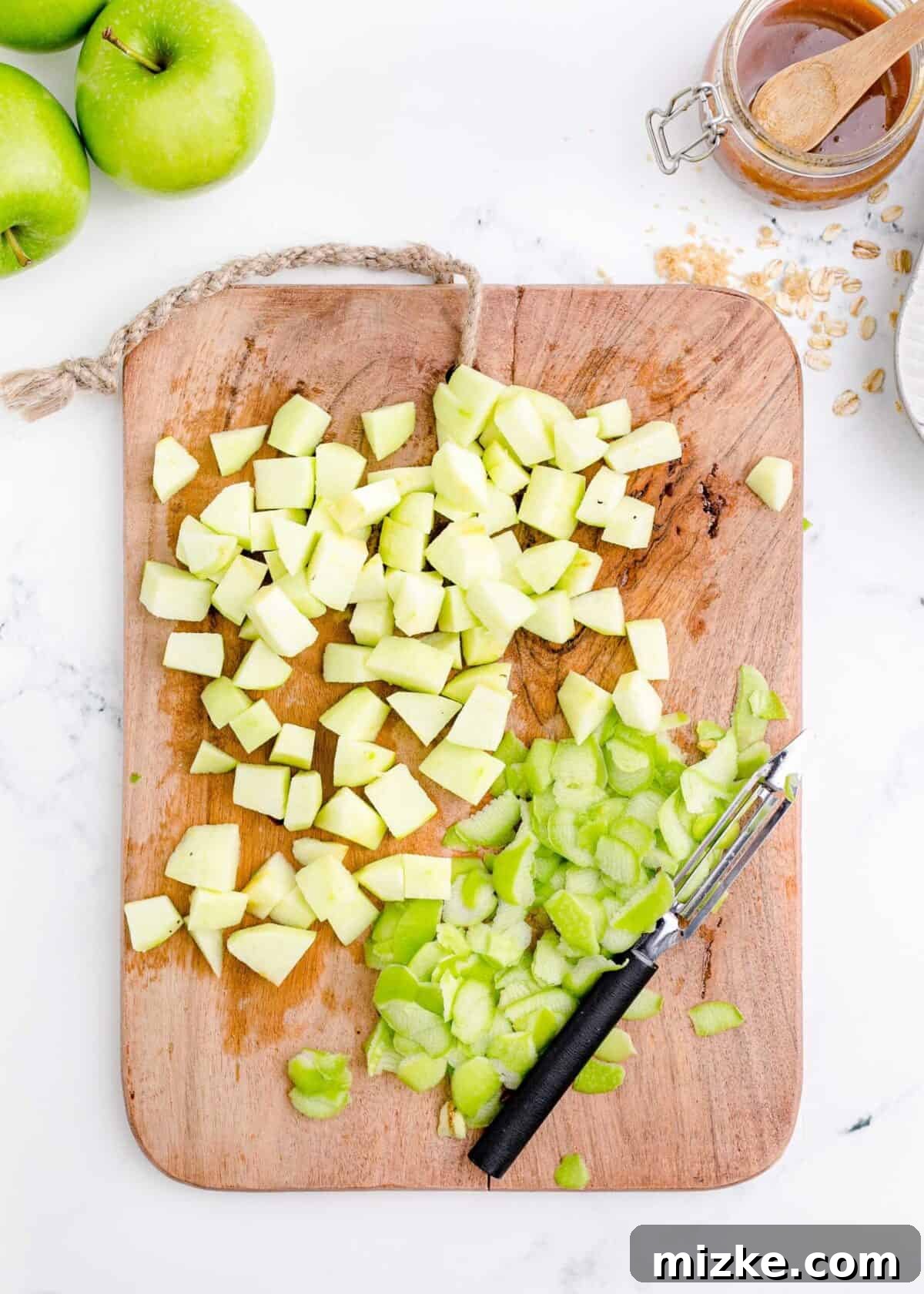 Diced apples on a cutting board, prepared for the cheesecake topping. A cutting board laden with finely diced apples, indicating careful preparation, with a peeler lying nearby and whole, untouched apples in the background, ready to be processed.