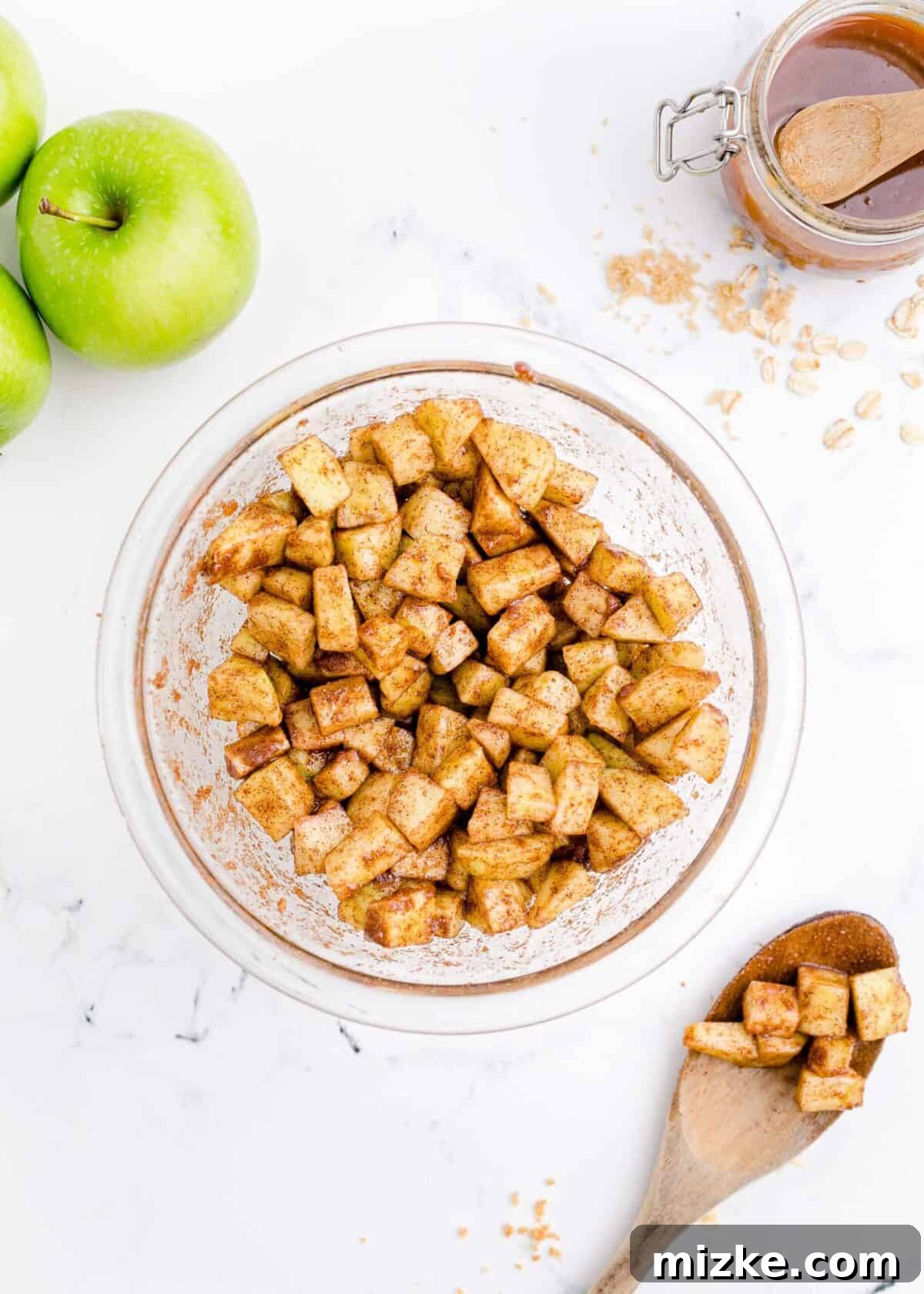 Mixing bowl of apples tossed with brown sugar and cinnamon. A mixing bowl filled with freshly diced apples, thoroughly coated in a glistening mixture of brown sugar and cinnamon, with a wooden spoon resting nearby, showing some of the sweet, spiced apples.