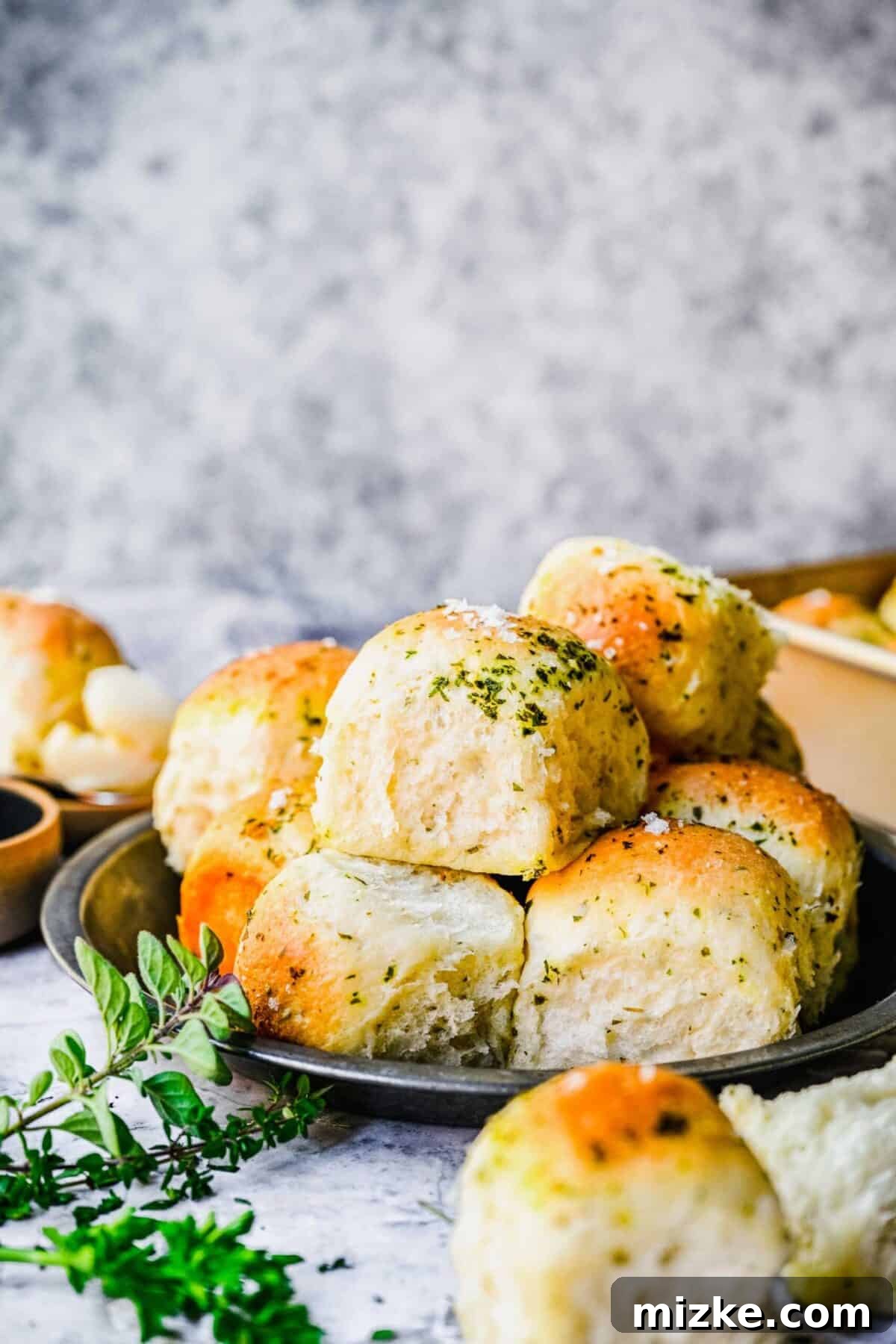 These easy garlic herb parker house rolls are buttery, moist, and tender, and the perfect side for a Thanksgiving or Christmas meal. A plate with a pile of golden-brown garlic herb parker house rolls, next to some fresh herbs, with more rolls in the foreground and background.
