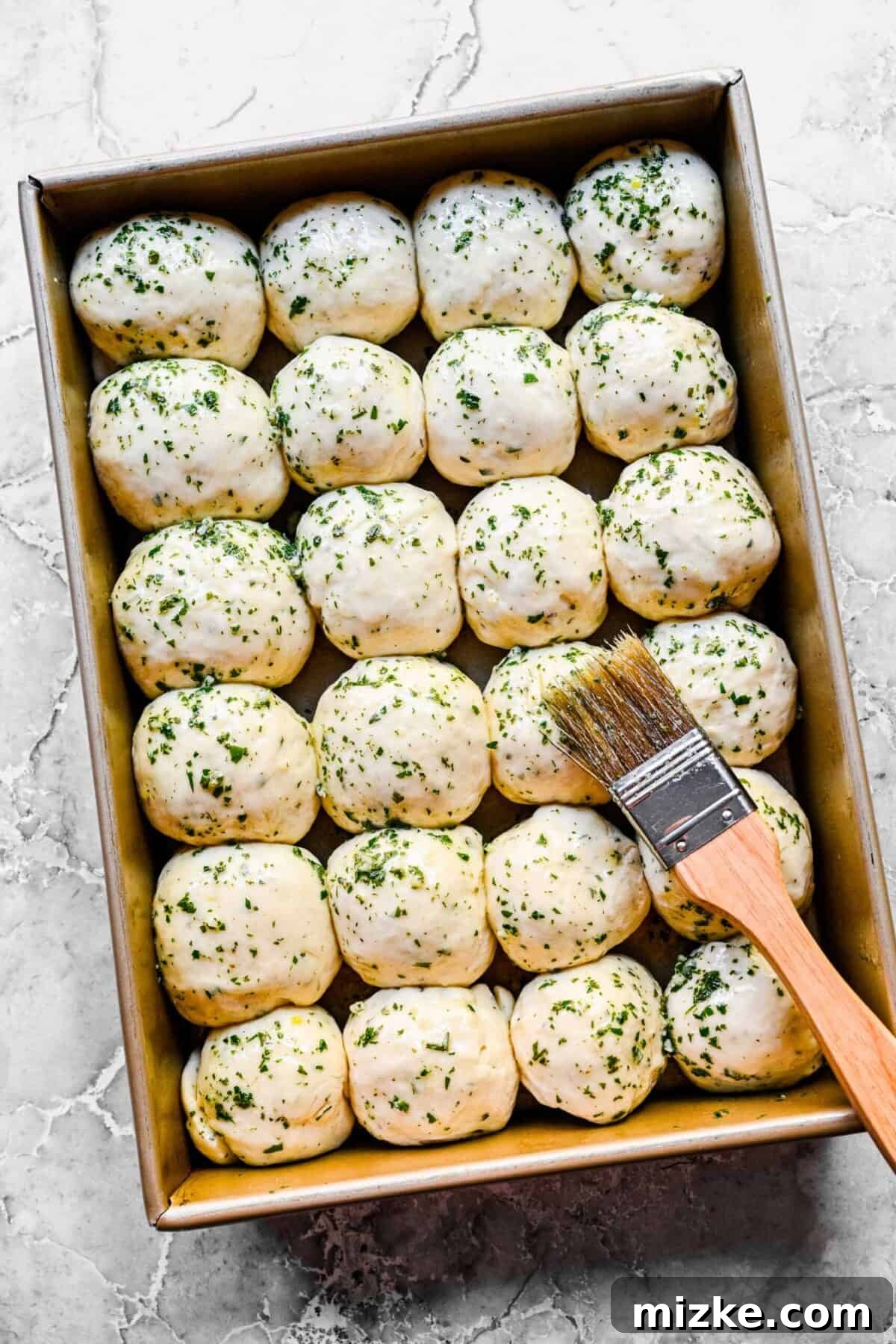 These easy garlic herb parker house rolls are buttery, moist, and tender, and the perfect side for a Thanksgiving or Christmas meal. A baking tray filled with 24 balls of uncooked garlic herb roll dough, lavishly covered in a butter, garlic, and herb mixture, with a baking brush resting on top.