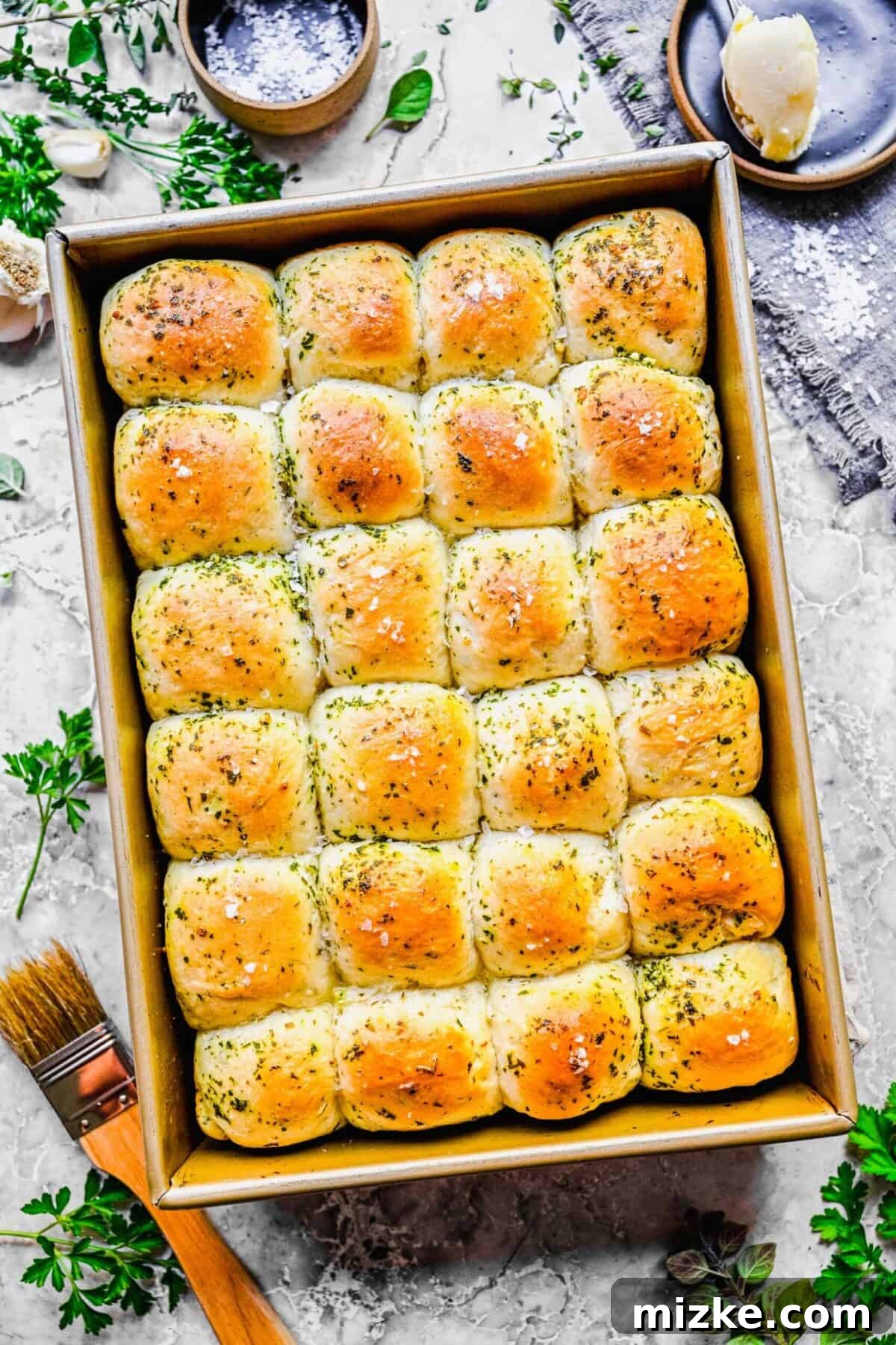 These easy garlic herb parker house rolls are buttery, moist, and tender, and the perfect side for a Thanksgiving or Christmas meal. A tray of 24 perfectly baked garlic herb parker house rolls, glistening with butter and topped with fresh herbs, positioned next to some fresh herbs and a baking brush.