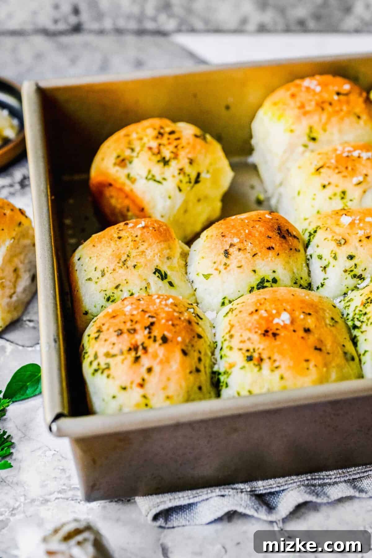 These easy garlic herb parker house rolls are buttery, moist, and tender, and the perfect side for a Thanksgiving or Christmas meal. The end of a baking tray filled with baked garlic herb parker house rolls, with a few rolls removed, showcasing their soft interior.