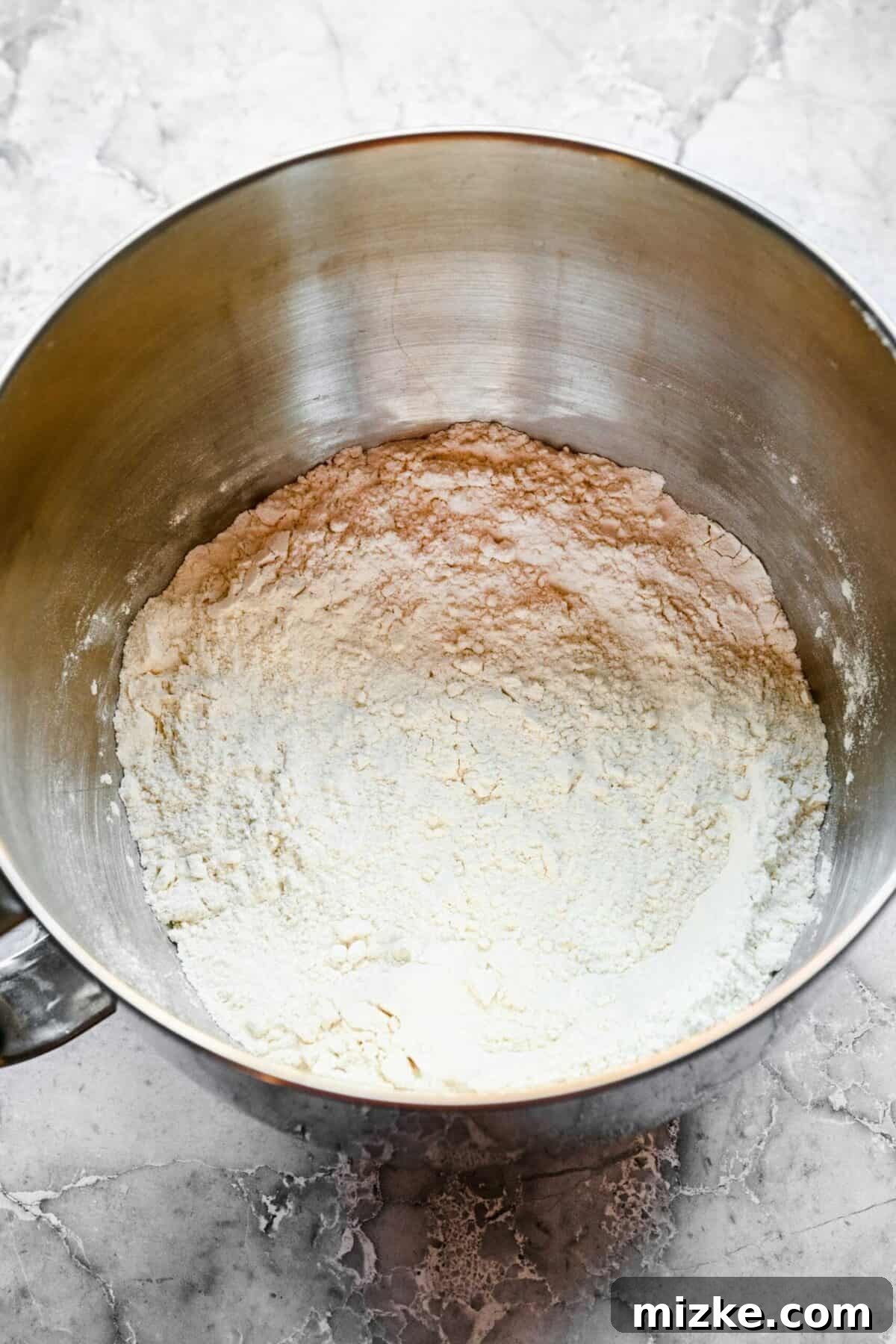 These easy garlic herb parker house rolls are buttery, moist, and tender, and the perfect side for a Thanksgiving or Christmas meal. A stand mixer mixing bowl filled with a flour mixture, ready for wet ingredients.