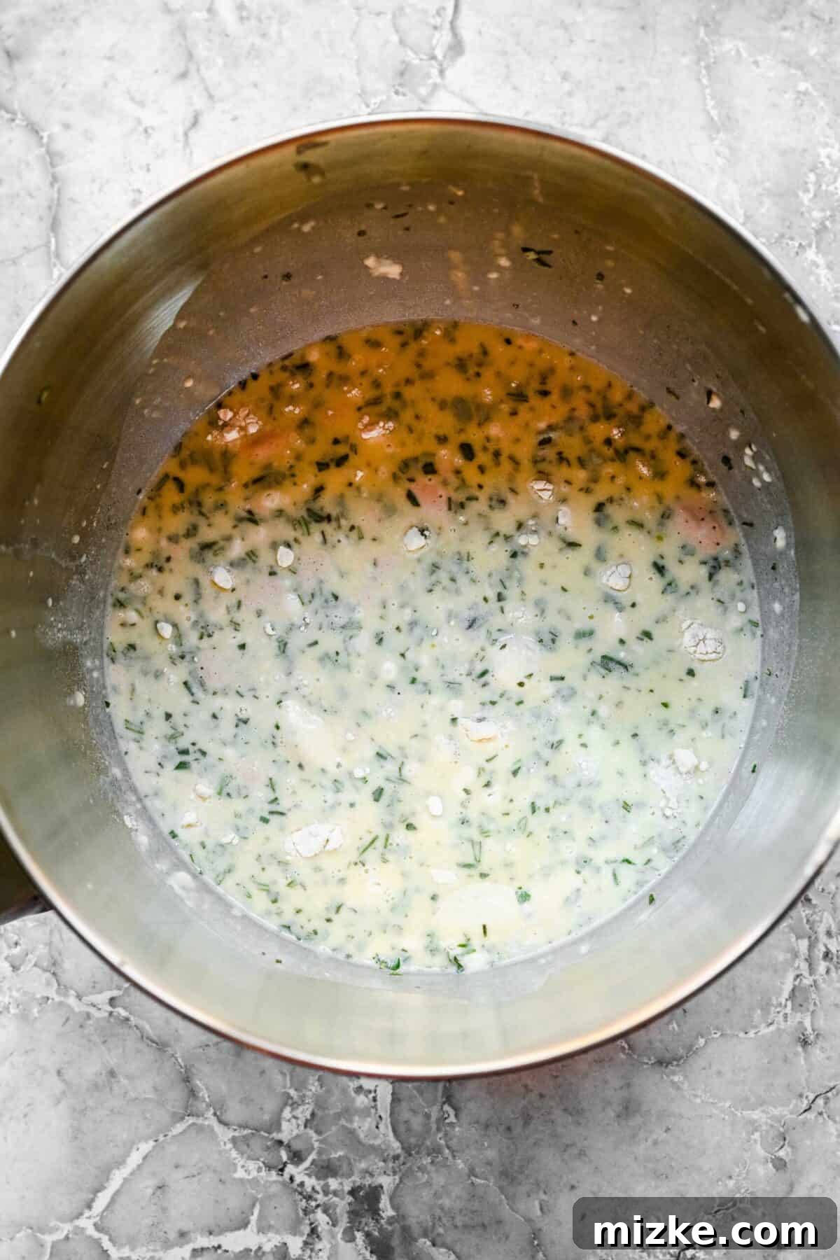 These easy garlic herb parker house rolls are buttery, moist, and tender, and the perfect side for a Thanksgiving or Christmas meal. A stand mixer mixing bowl with a flour mixture, with a milk and butter mixture poured on top, brimming with garlic and herbs.