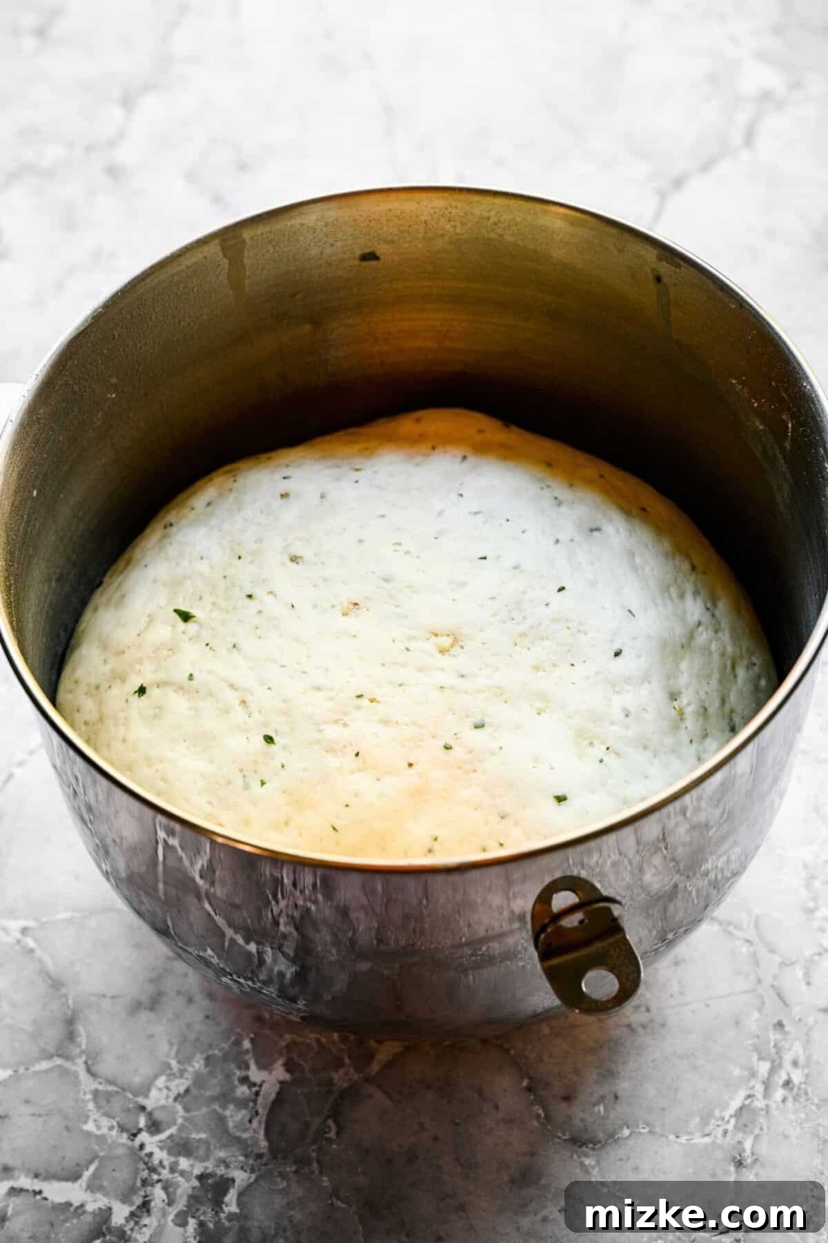 These easy garlic herb parker house rolls are buttery, moist, and tender, and the perfect side for a Thanksgiving or Christmas meal. A stand mixer bowl filled with puffy, risen garlic herb roll dough, showing its doubled volume.