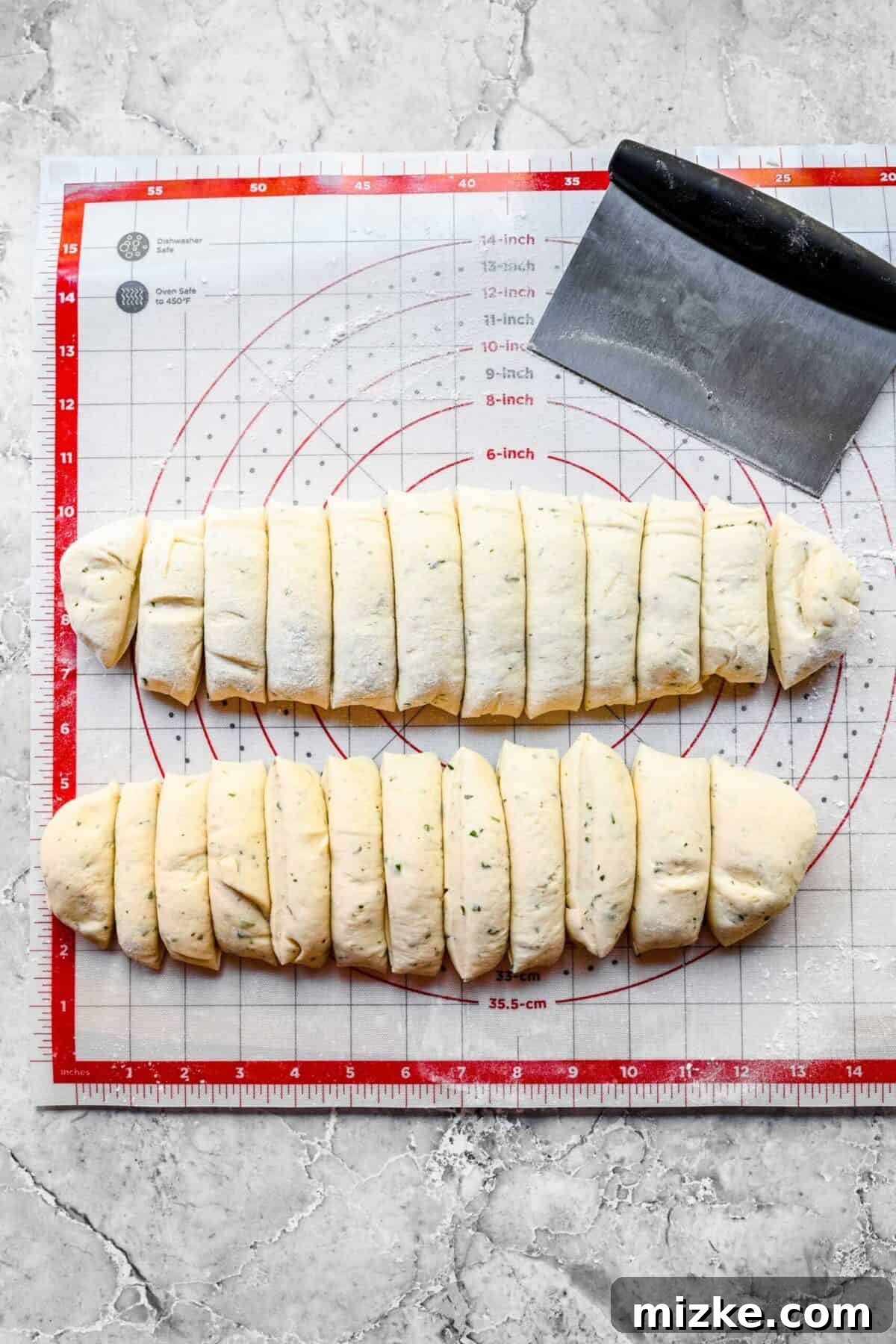 These easy garlic herb parker house rolls are buttery, moist, and tender, and the perfect side for a Thanksgiving or Christmas meal. Two logs of garlic herb parker house roll dough, each precisely cut into 12 pieces, arranged on a silicone mat with measurements, alongside a bench scraper.