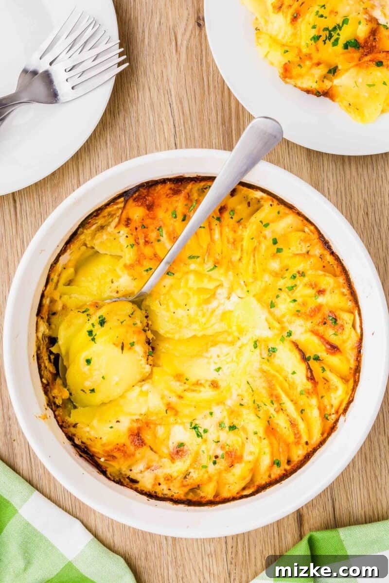 Simply Irresistible Creamy Potato Gratin 2 An overhead shot showcasing a rustic wooden cutting board alongside a white casserole dish filled with golden-brown potatoes au gratin, with a silver spoon poised to serve a scoop.
