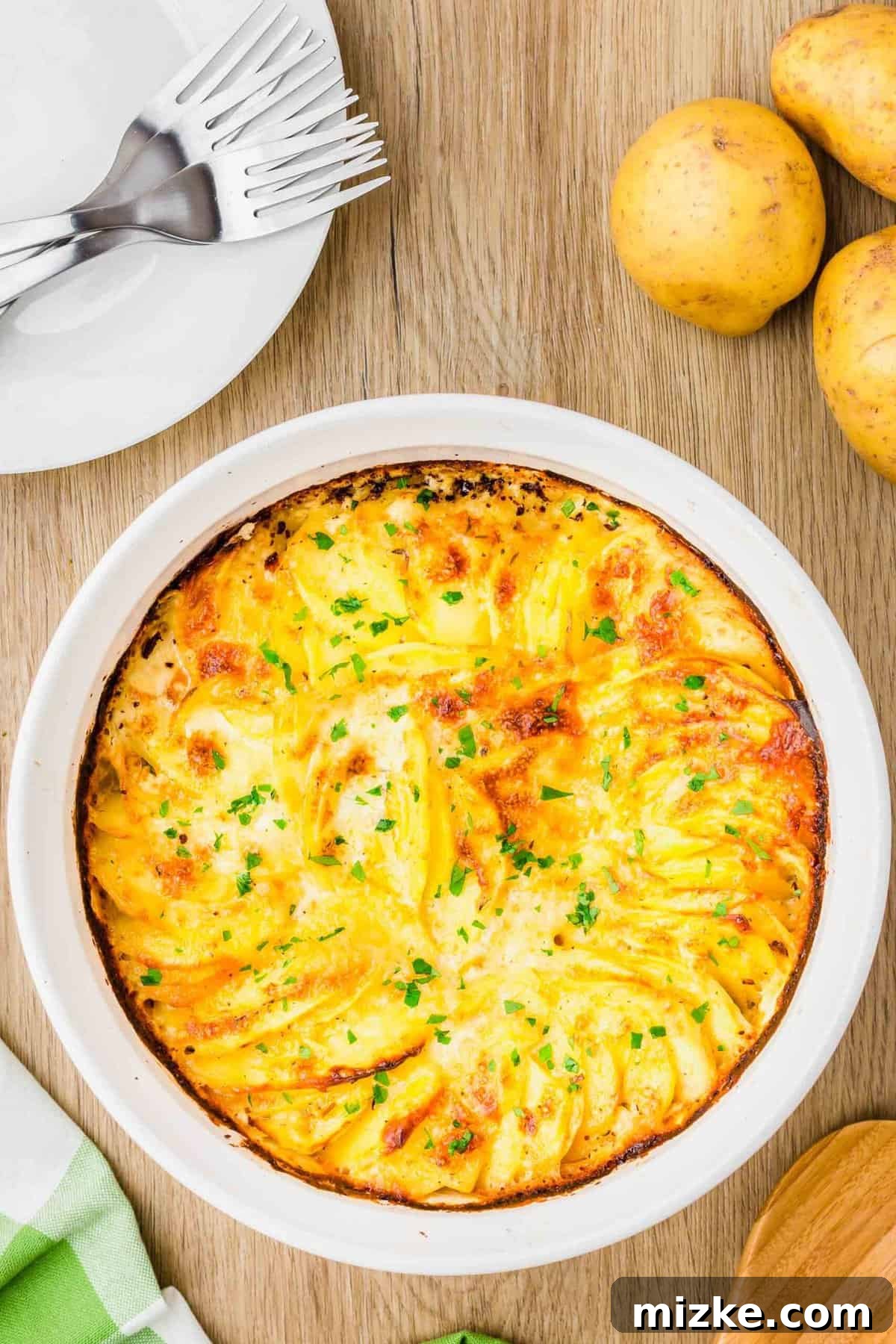Simply Irresistible Creamy Potato Gratin 11 An inviting overhead shot of baked potatoes au gratin, fresh from the oven, garnished with vibrant fresh parsley in a casserole dish. The dish rests on a wooden cutting board beside a plaid green towel, a wooden spoon, and a plate with silver forks, setting a warm, rustic scene.