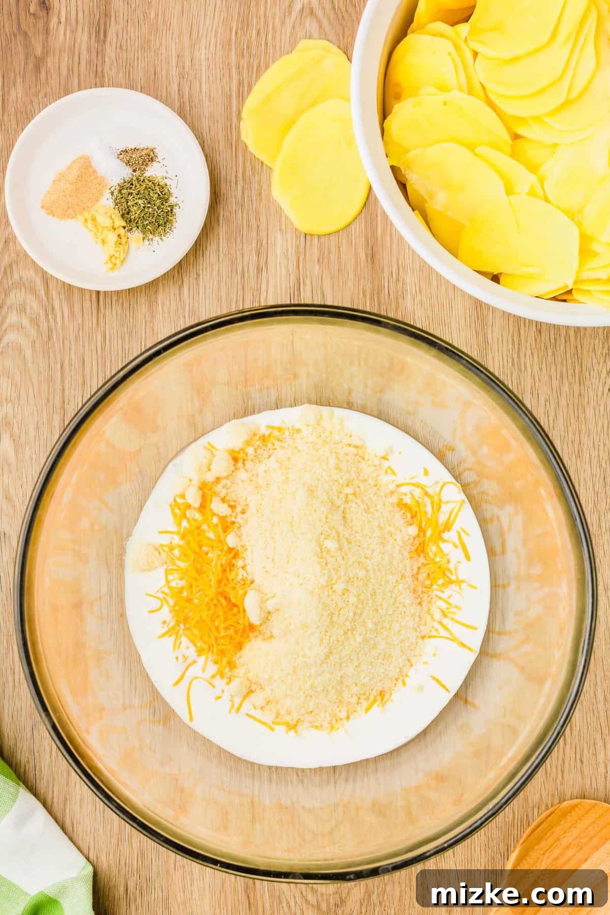 Simply Irresistible Creamy Potato Gratin 6 A clear mixing bowl filled with heavy cream and cheeses, positioned next to a small plate of spice seasonings and a pile of freshly sliced yellow potatoes, ready for mixing.