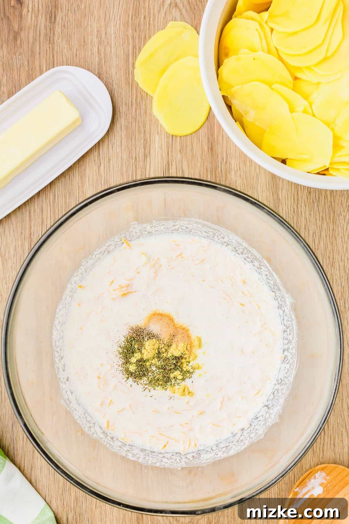 Simply Irresistible Creamy Potato Gratin 7 Inside a clear mixing bowl, heavy cream, cheddar, and parmesan cheeses are thoroughly combined, with spice seasonings already incorporated, flanked by a stick of butter and a stack of sliced potatoes.