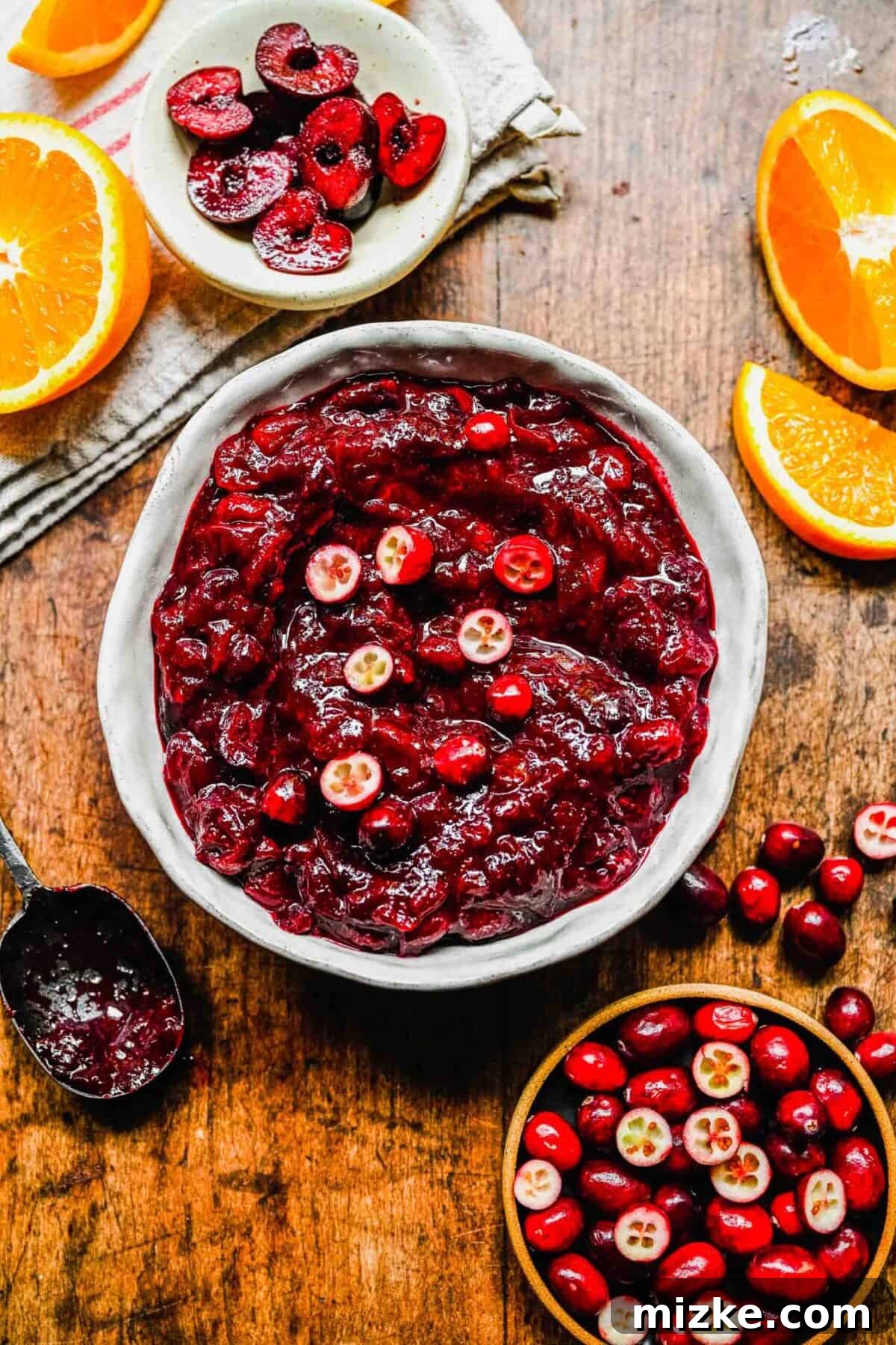 This cranberry cherry compote recipe is tart and sweet, takes only 25 minutes to make, and can be made in advance for any Thanksgiving meal. A bowl of cranberry cherry compote topped with fresh cranberries, next to a bowl of cranberries, a bowl of cherries, a dirty spoon, and some orange slices