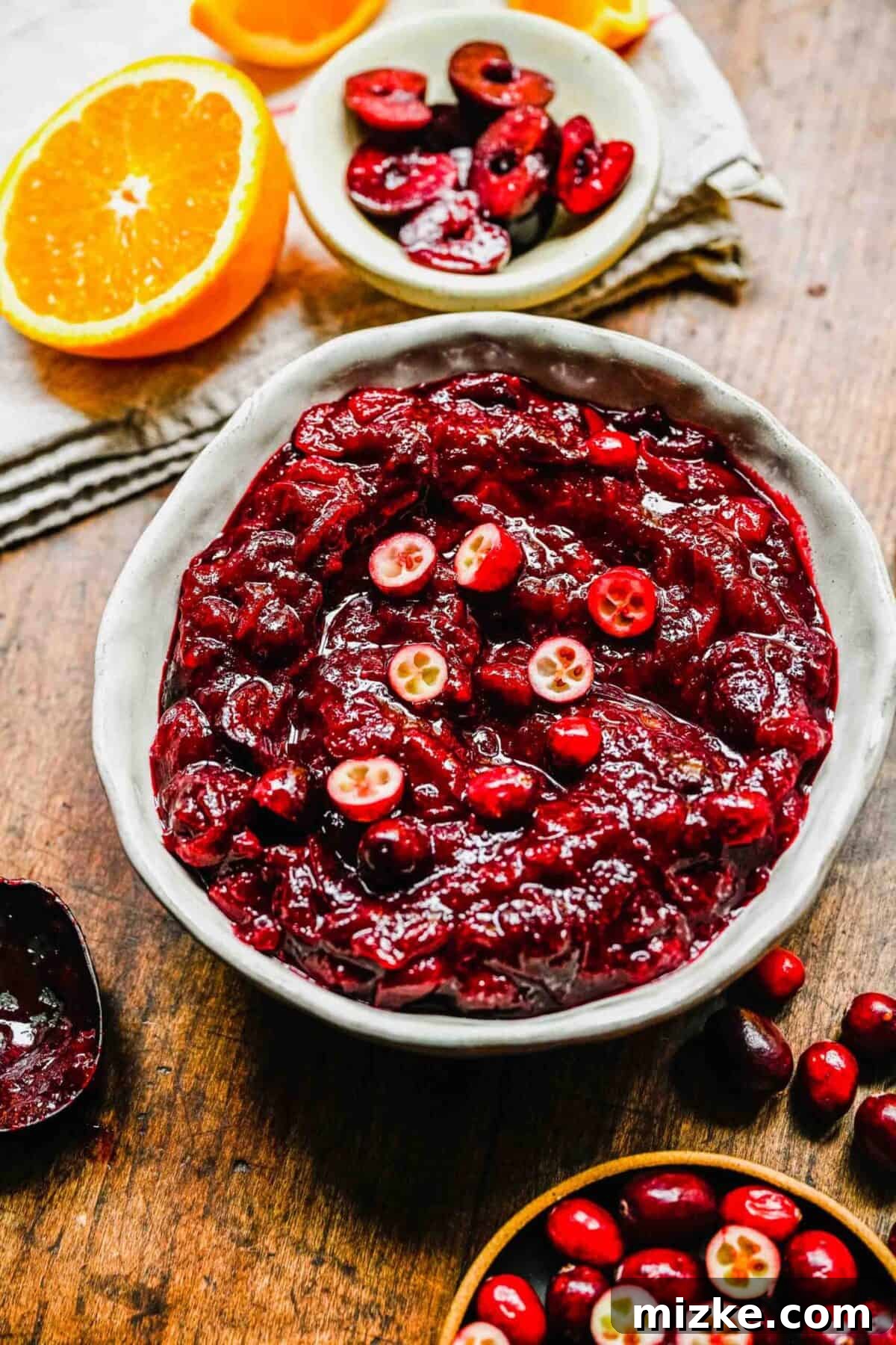 This cranberry cherry compote recipe is tart and sweet, takes only 25 minutes to make, and can be made in advance for any Thanksgiving meal. A bowl of cranberry cherry compote topped with sliced cranberries, next to half an orange, a bowl of cranberries, a bowl of cherries, and a dirty spoon